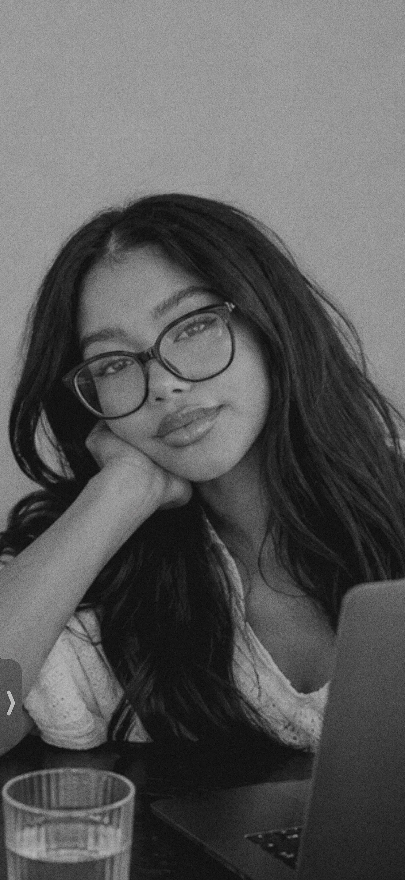 A young woman with long dark hair and glasses, resting her head on her hand, looking at the camera with a slight smile in a black-and-white photo.