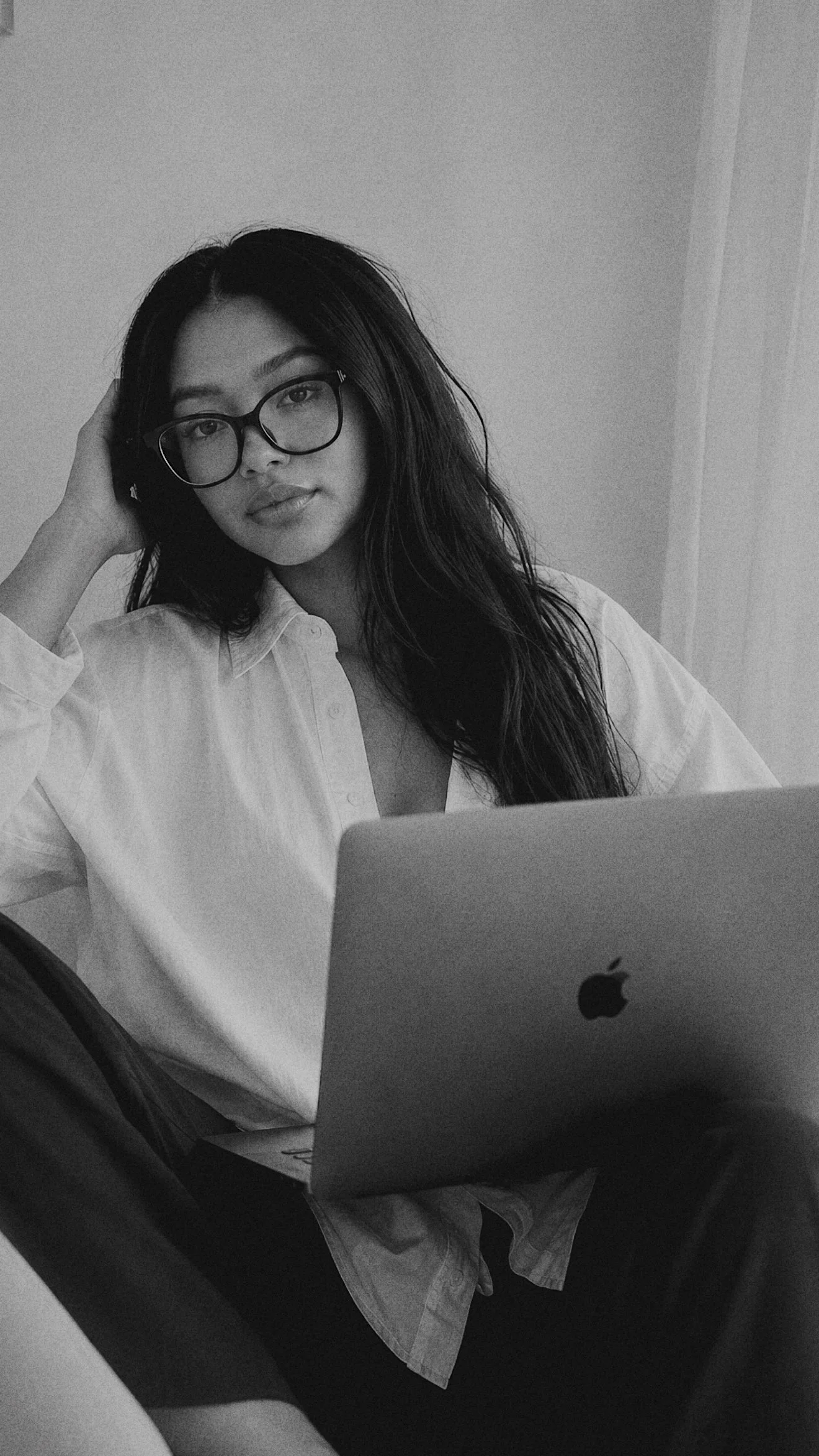 A woman with long dark hair and glasses wearing a white shirt, sitting at a desk with a laptop and looking at the camera in a black-and-white photo.