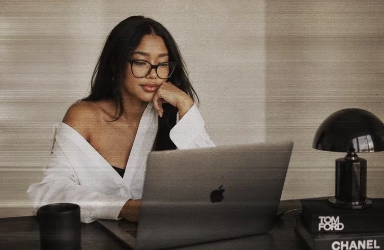 A woman with long dark hair and glasses working on a laptop at a desk, with a black desk lamp, a black cup, and books including one with 'CHANEL' and another with 'TOM FORD' on it.
