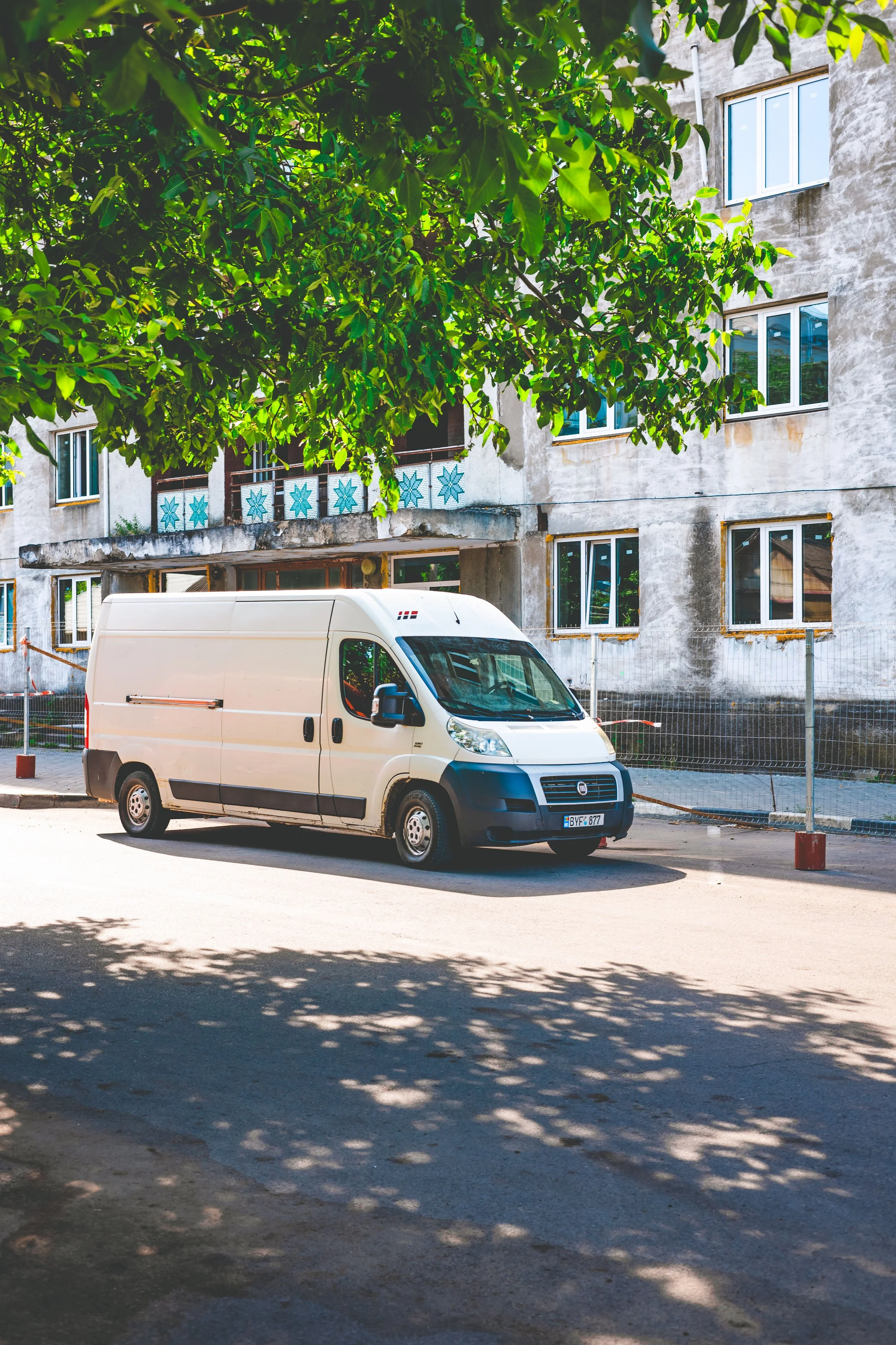 Leere weiße Lieferwagen parkt auf der Straße vor einem grauen, älteren Wohngebäude mit mehreren Fenstern. Ein großer Baum wirft Schatten auf die Straße.