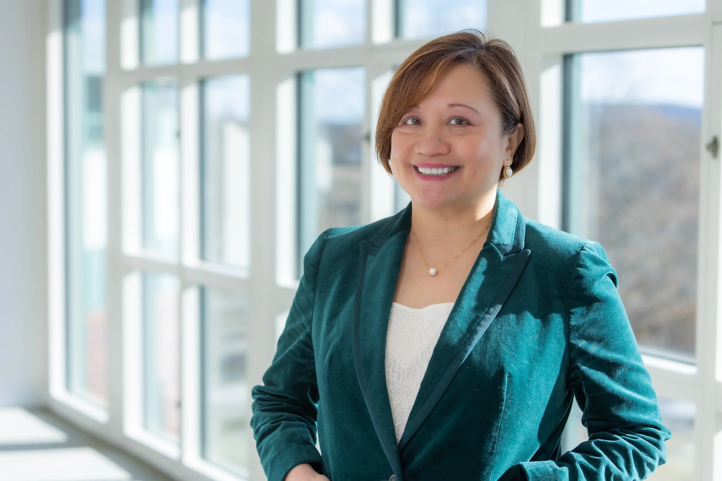 Smiling woman with short brown hair wearing a teal blazer and jewelry, standing in front of large windows in a bright, modern indoor space.
