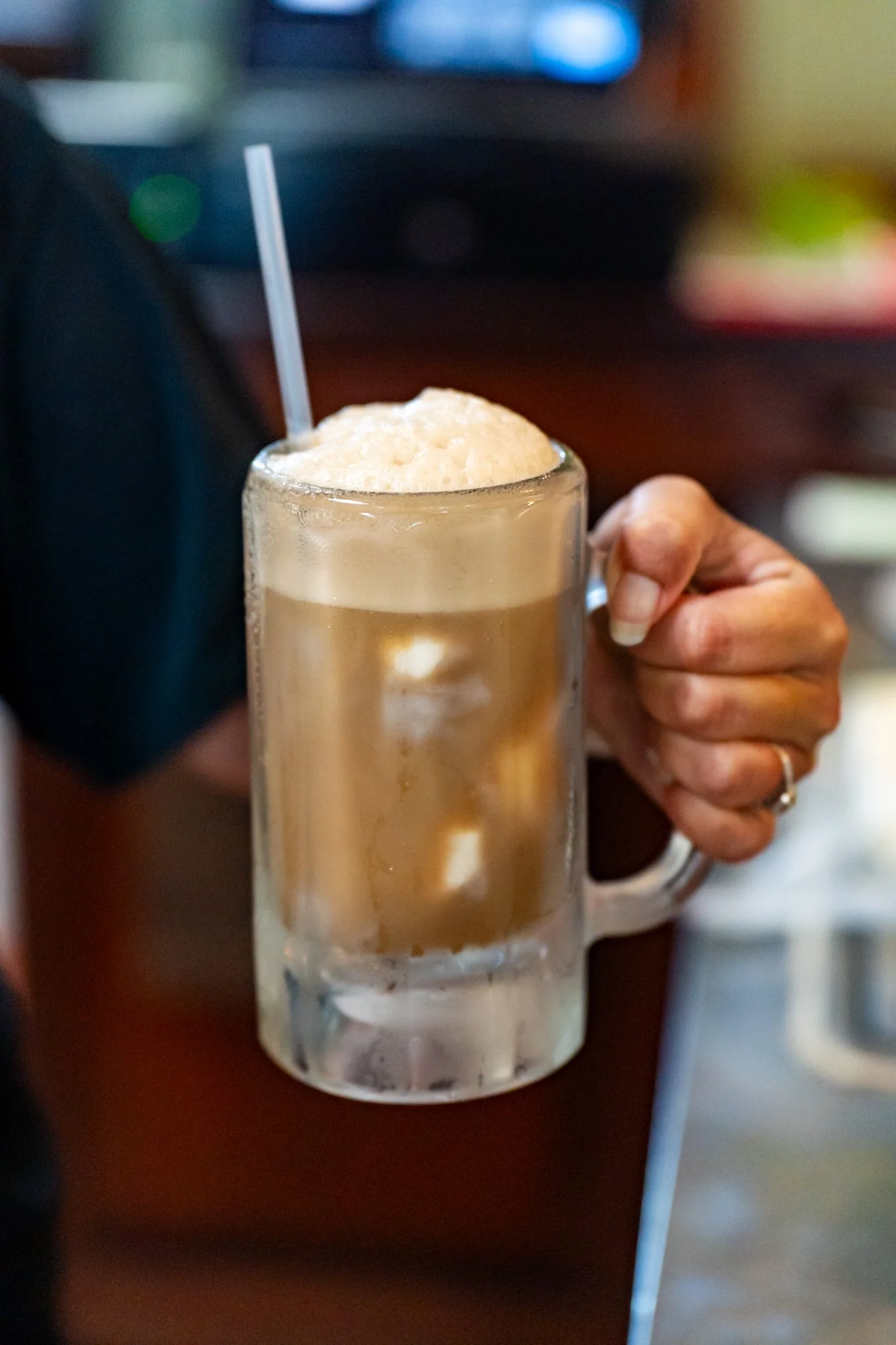 A hand holding a clear glass mug filled with a topped beverage, possibly coffee or latte, with ice and a straw, on a dark wooden surface.
