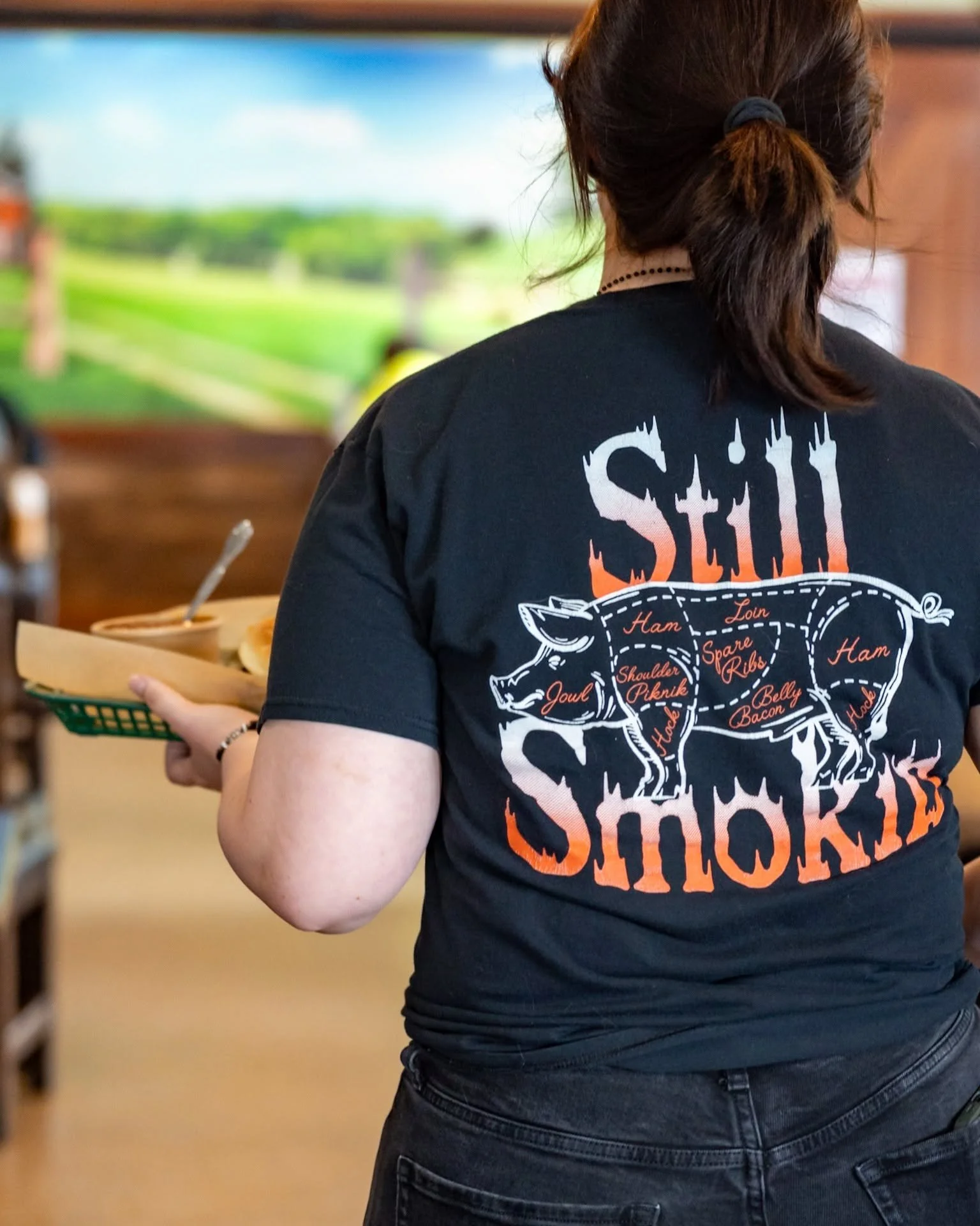A woman with brown hair tied back in a ponytail, wearing a black T-shirt with a pig diagram and the word 'SHOCK' on the back, is holding a tray of food in a casual restaurant or cafe setting.