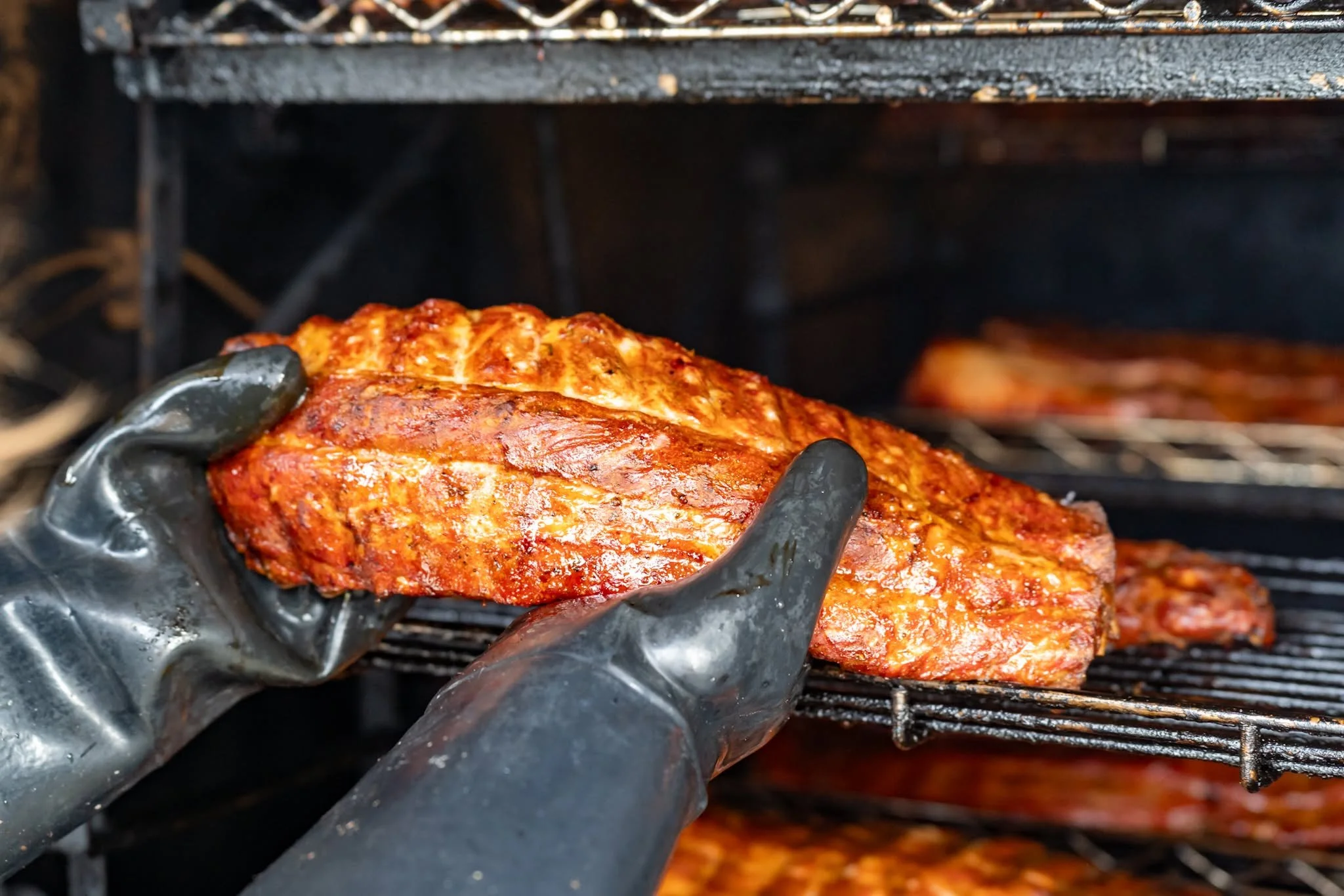 Hand wearing black gloves holding a rack of barbecue ribs with a smoky grill in the background.