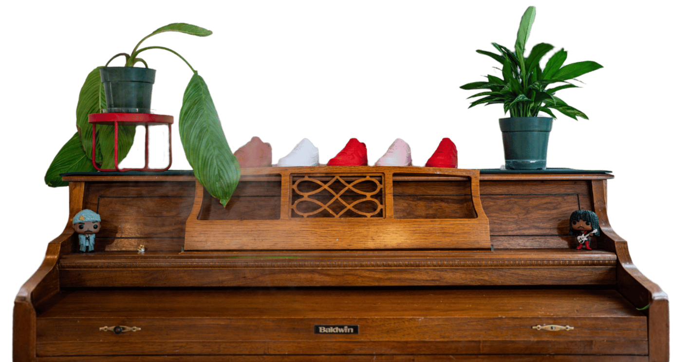 A wooden Baldwin piano with two potted plants on top, one on each side, and five shoes in between. Two small figurines are on each side of the piano, one of a person with dark hair and the other with a light blue outfit.