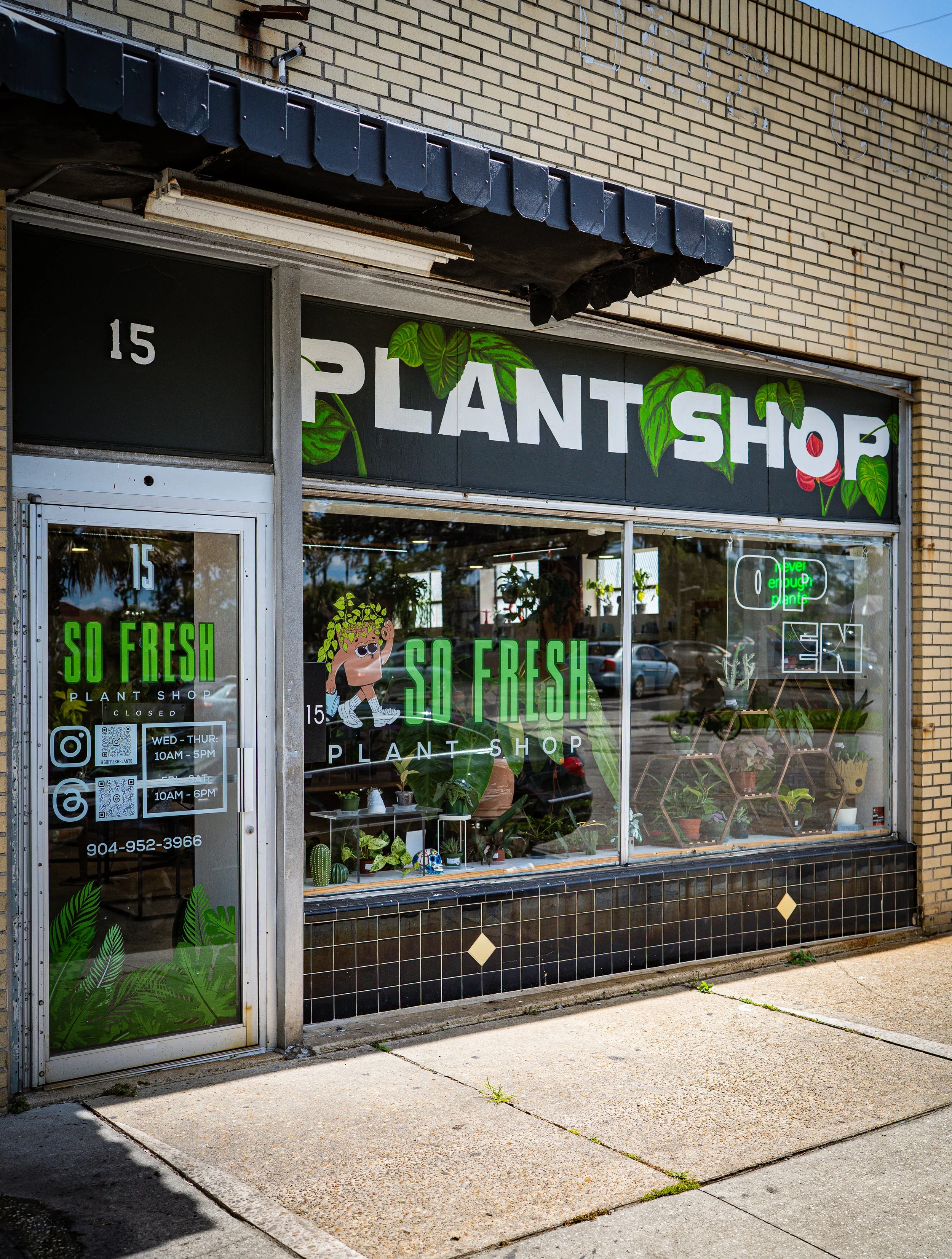 Front view of a plant shop with large windows, decorated with green leaves and a cartoon character, displaying the text 'PLANT SHOP' on the signage and window. The shop's address is 15, and the shop hours are Wednesday to Thursday from 10AM to 5PM and Saturday from 10AM to 6PM. A neon sign inside the shop reads 'open.' There is a reflection of cars and trees in the window, and the sidewalk is concrete with some patches of grass.