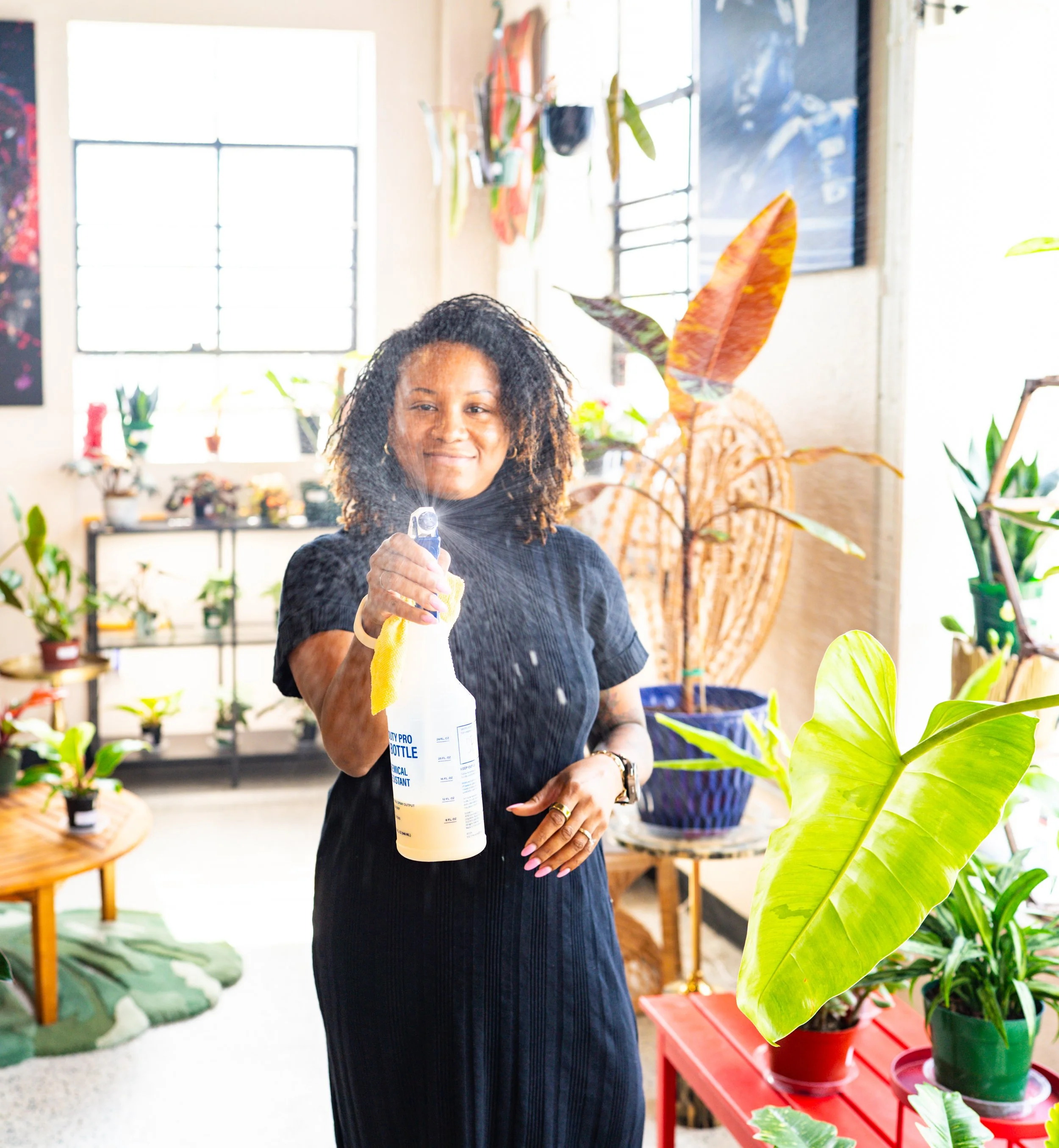 A woman with curly hair, wearing a black dress, is in a room filled with plants. She is holding a spray bottle and a yellow cloth, smiling slightly and spritzing water towards the viewer. Potted plants and a colorful decor in the background.