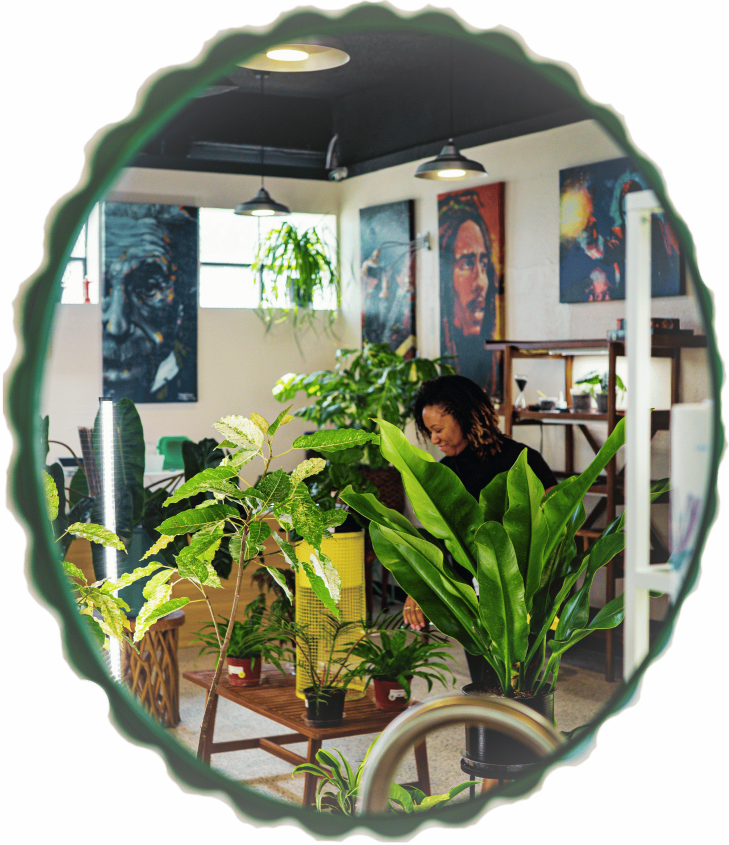 A woman with curly hair working in a room filled with various green plants. The room has artwork on the walls, including portraits, and is lit by hanging lights. The photo is taken through a mirror with a wavy green edge.