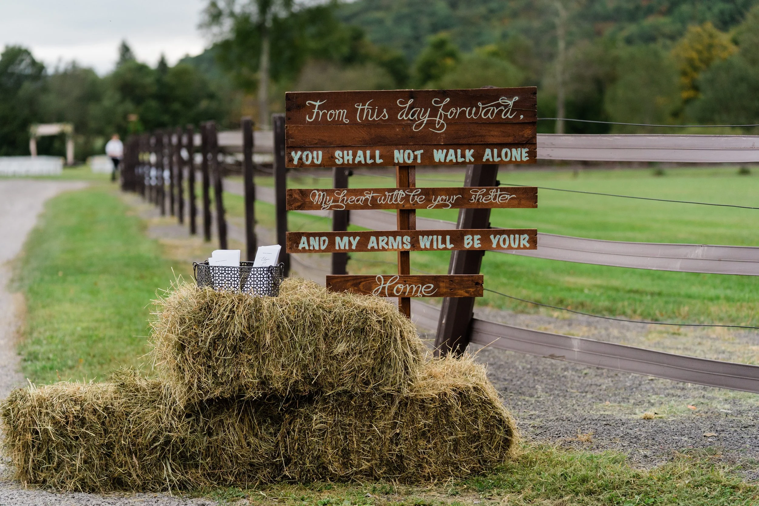 A rustic wooden sign with white and blue lettering that reads, "From this day forward, you shall not walk alone. My heart will be your shelter and my arms will be your home." The sign is leaning against a hay bale near a fence in a rural outdoor sett