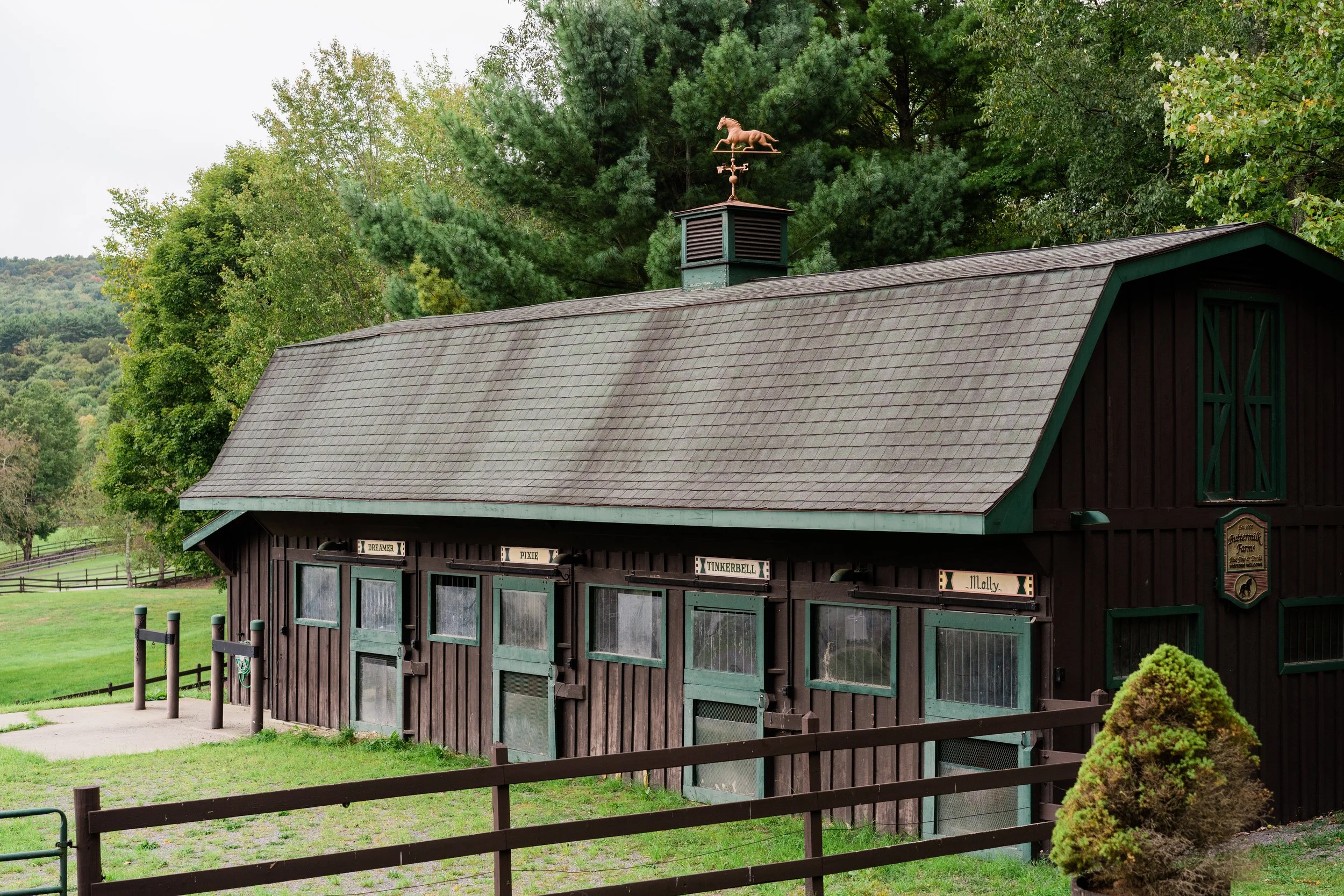A wooden barn with a shingled roof surrounded by trees and a grassy area, with a weather vane on top of the barn featuring a horse silhouette.