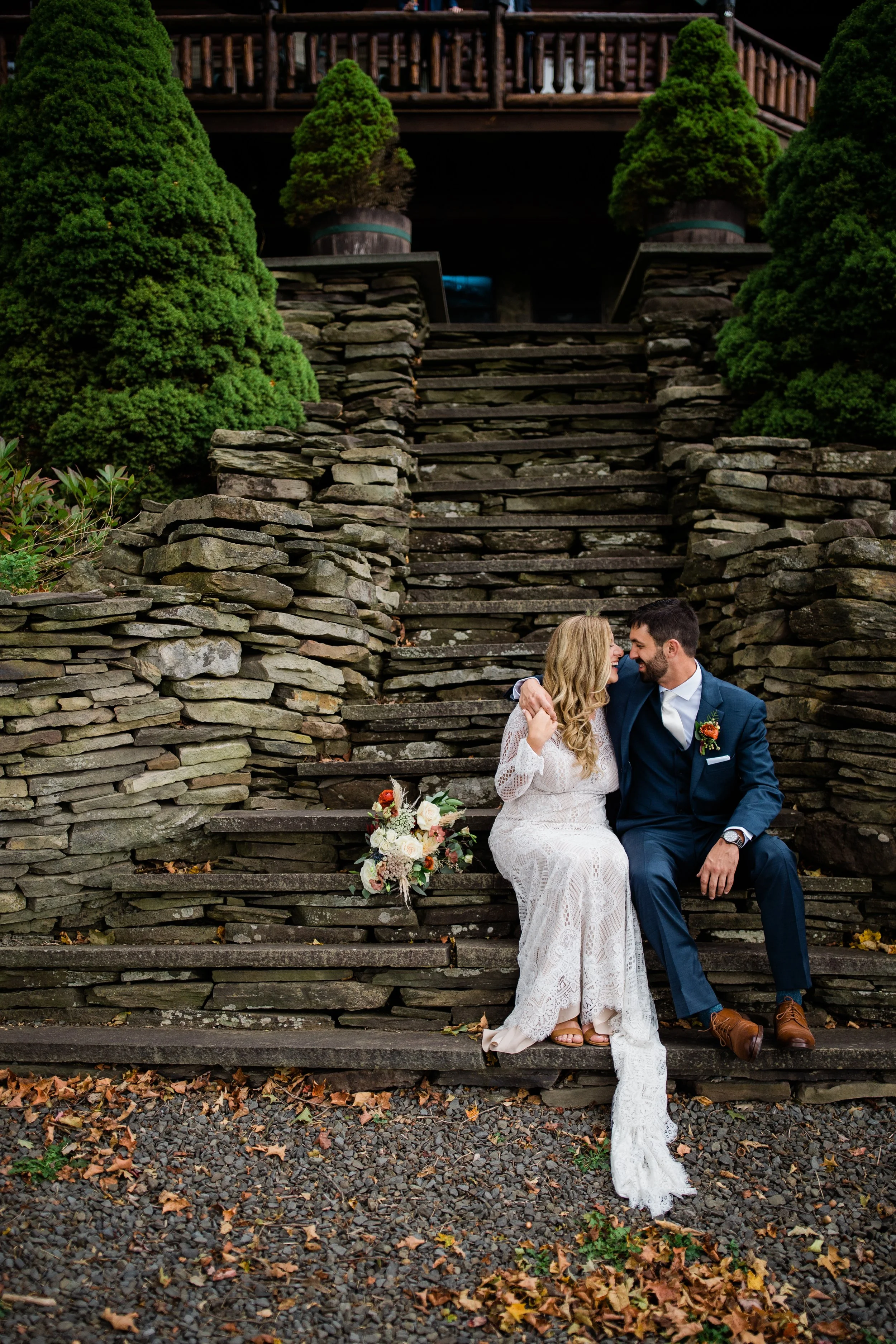 A newlywed couple sits on stone steps outdoors, embracing and smiling at each other, with a bridal bouquet placed beside them, surrounded by fallen autumn leaves and lush green trees.