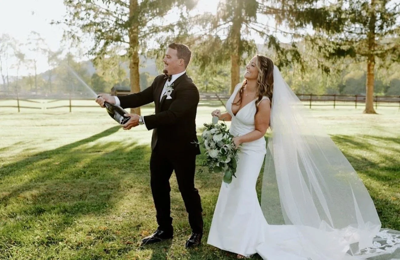 A bride and groom celebrating outdoors, with the groom opening a champagne bottle that is spraying, while the bride holds a bouquet of flowers and smiles.