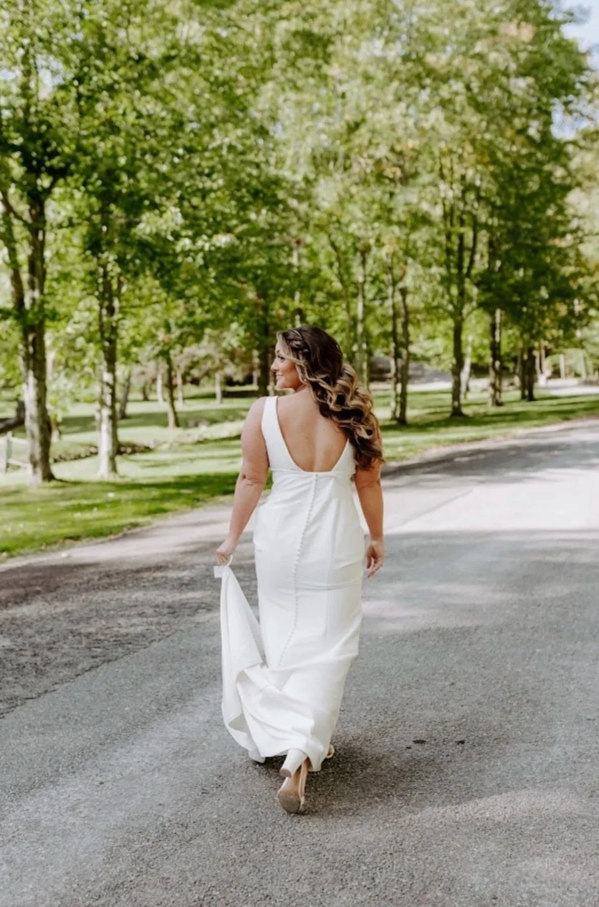 A woman in a white gown walking on a paved path in a park with green trees.