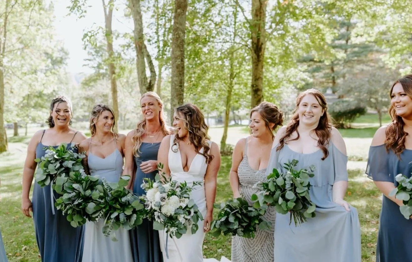 A group of bridesmaids and a bride at a wedding outdoors, holding bouquets, surrounded by trees, laughing and smiling.