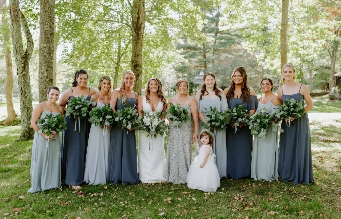 A bride and nine bridesmaids, along with a flower girl, posing outdoors in a park with trees. The women are dressed in shades of gray and holding bouquets of greenery, with the bride in a white dress.