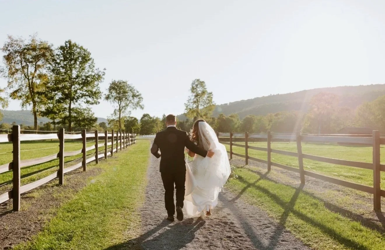 A bride and groom walking on a dirt path in a rural outdoor setting during sunset, with trees and distant hills in the background.