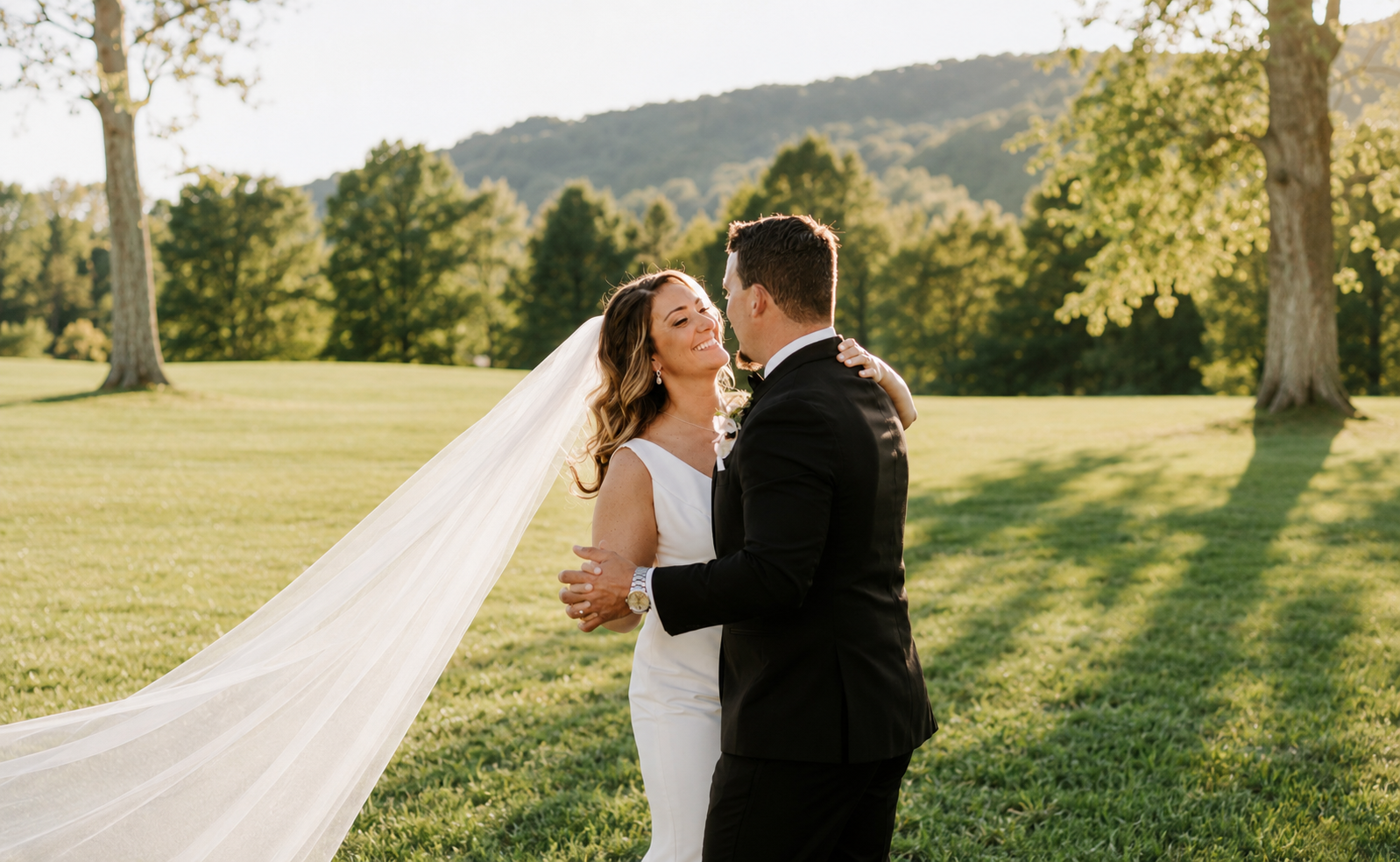 A bride and groom dancing outdoors on a sunny day with green grass, trees, and hills in the background.