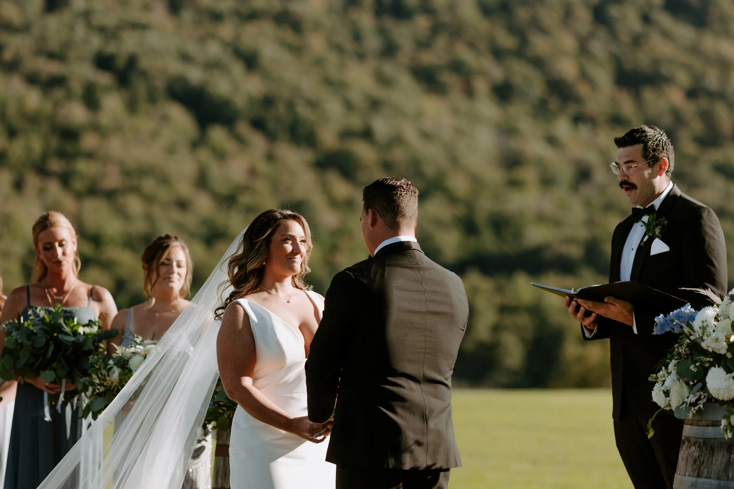 A wedding ceremony outdoors with a bride and groom holding hands, facing each other, with the officiant nearby. Four bridesmaids stand behind the bride, holding bouquets. The background features a green, hillside landscape.
