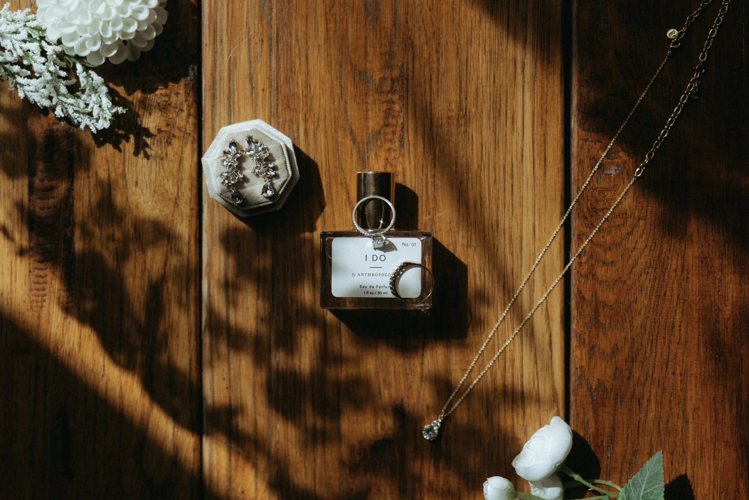 Jewelry and perfume on a wooden surface, including earrings, a necklace, a ring, and a perfume bottle, with flowers nearby.