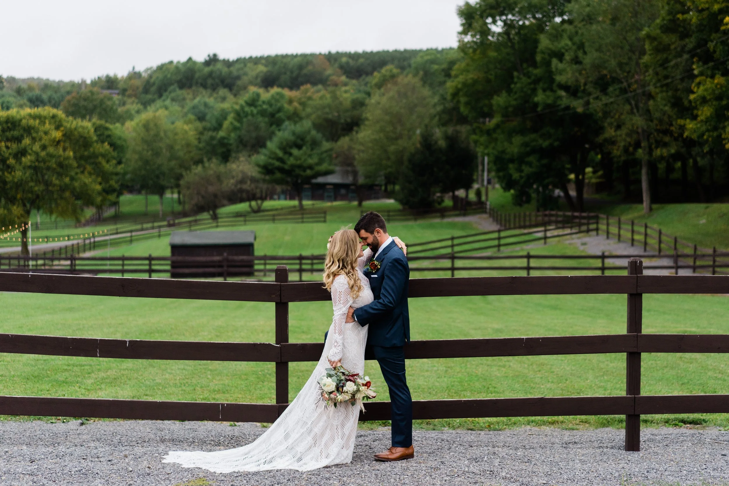 A bride and groom standing close together outdoors, smiling and looking at each other, in front of a wooden fence with a lush green landscape and trees in the background.