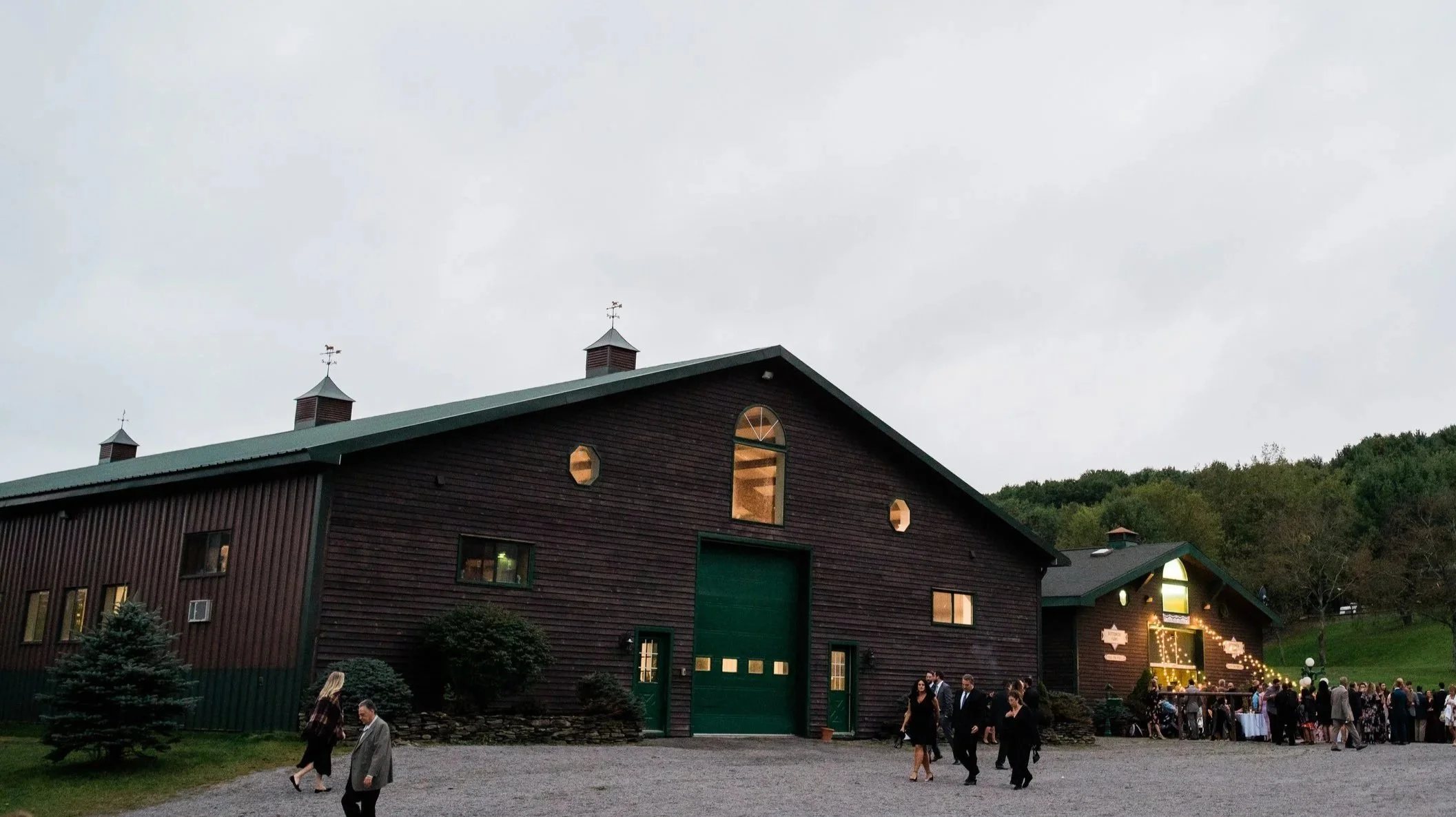 The exterior of a dark wooden barn-style building with a green garage door, multiple windows, and three small weather vanes on the roof, set against a backdrop of trees and a cloudy sky. People are gathered outside, some walking and some in small groups, near a well-lit reception area with string lights.