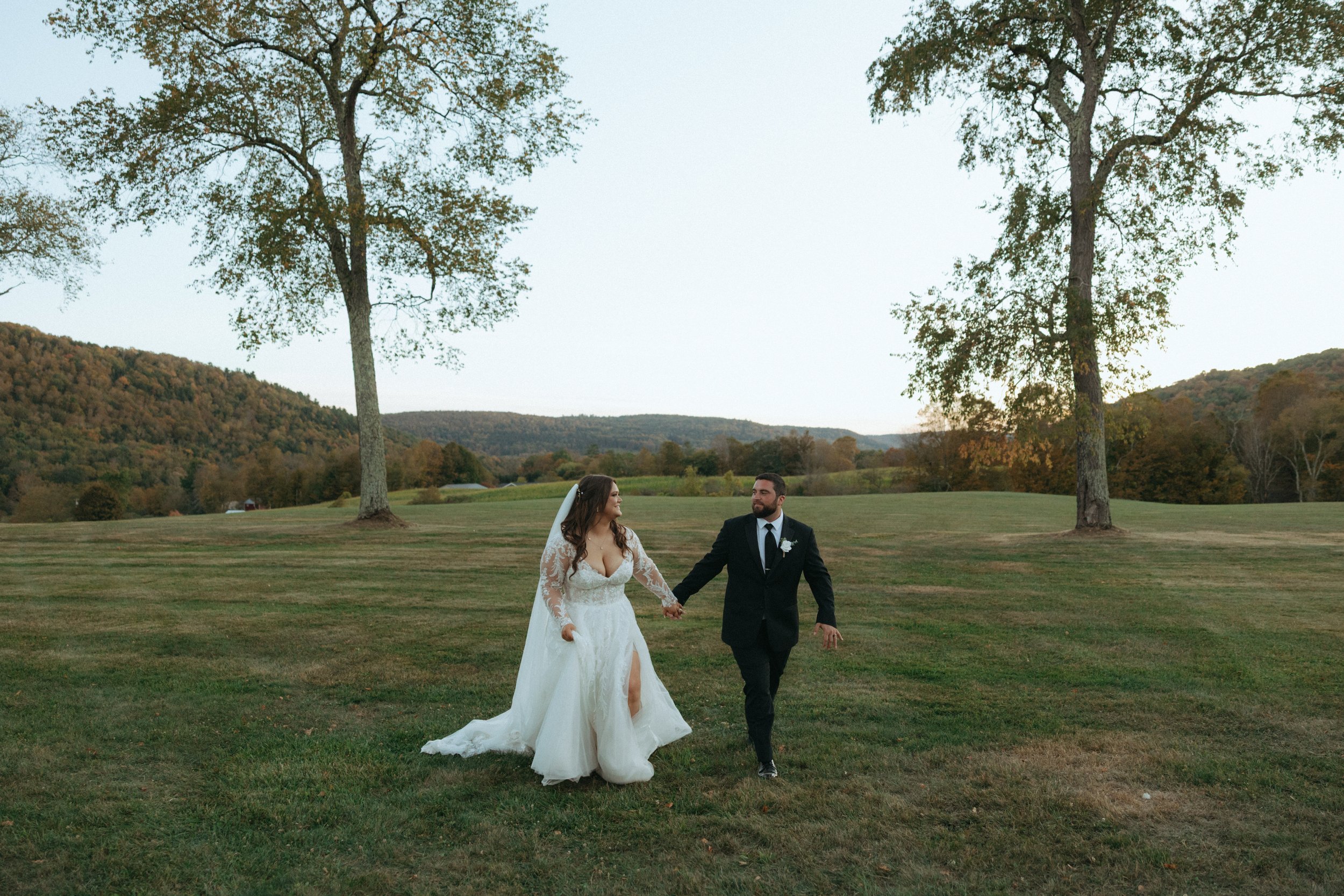A bride and groom holding hands and walking across a grassy field during their wedding ceremony outdoors, with trees and hills in the background, in the late afternoon or early evening.