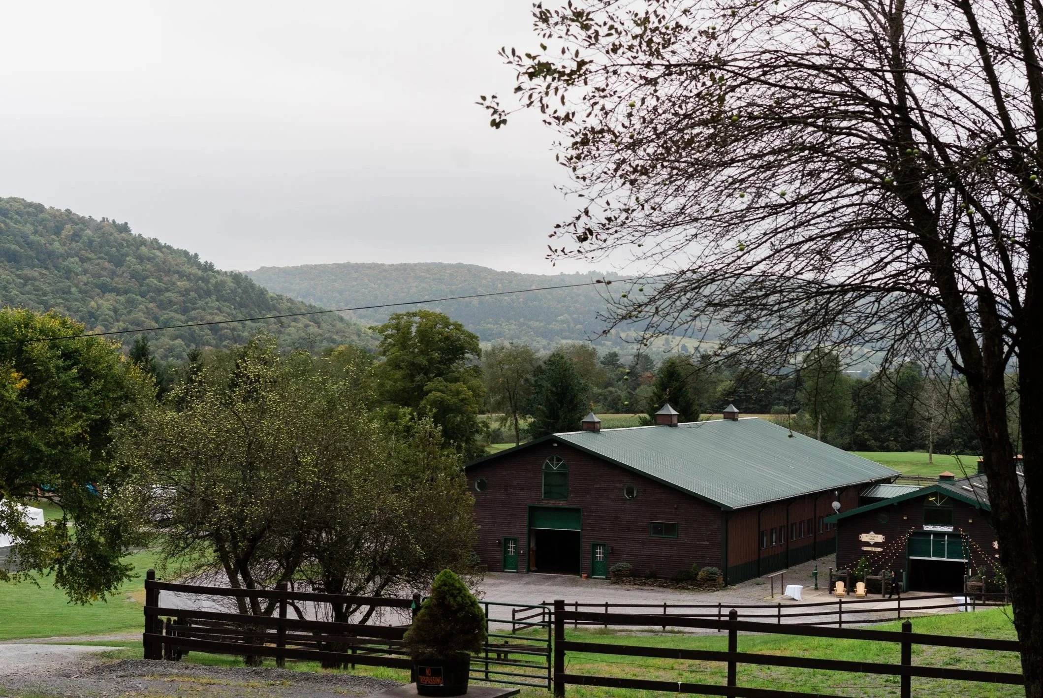 View of a rural landscape with rolling hills covered in trees under a cloudy sky. In the foreground, there is a wooden fence, a tree with sparse leaves and some green fruits, and a large barn with a metal roof and green doors. Nearby buildings include small structures with similar roofs. The scene has a peaceful, countryside atmosphere.