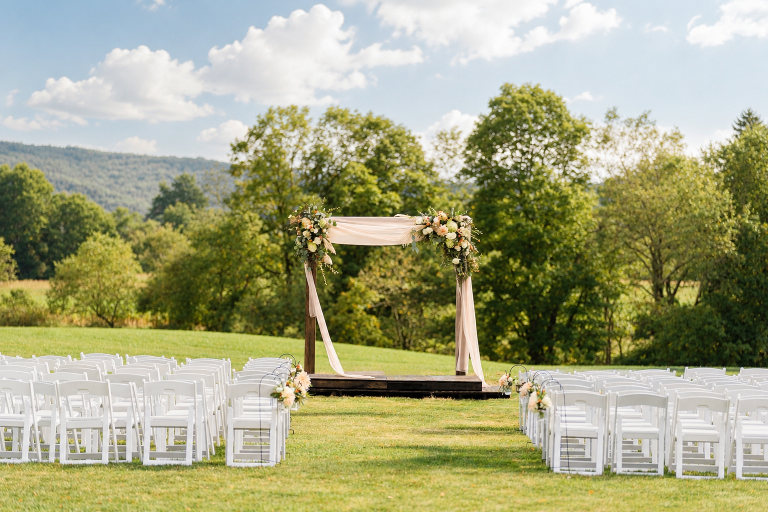 Outdoor wedding ceremony setup on a grassy field with white chairs arranged in rows facing a decorated wooden arch with flowers and fabric, surrounded by green trees and mountains under a partly cloudy sky.