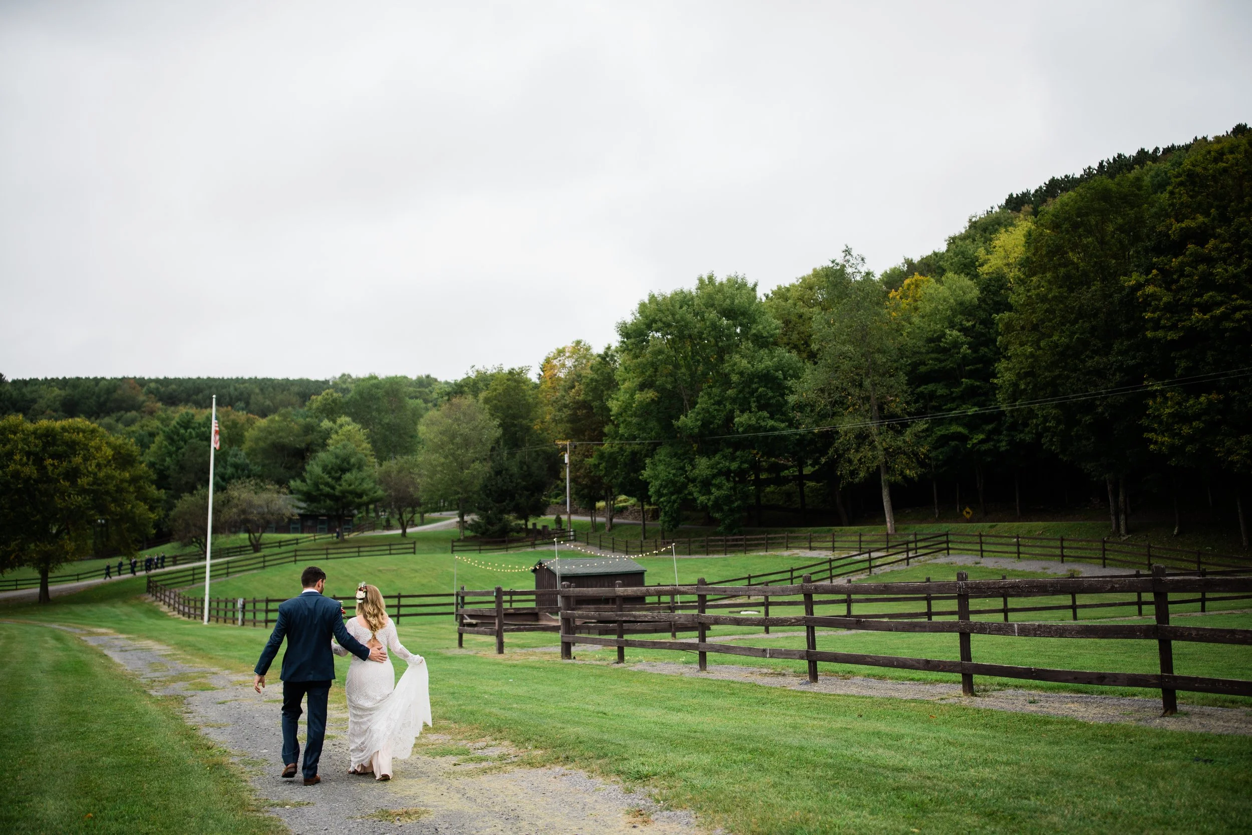 A bride and groom walking together on a grassy path at an outdoor wedding venue surrounded by a wooden fence, trees, and hills in the background on a cloudy day.