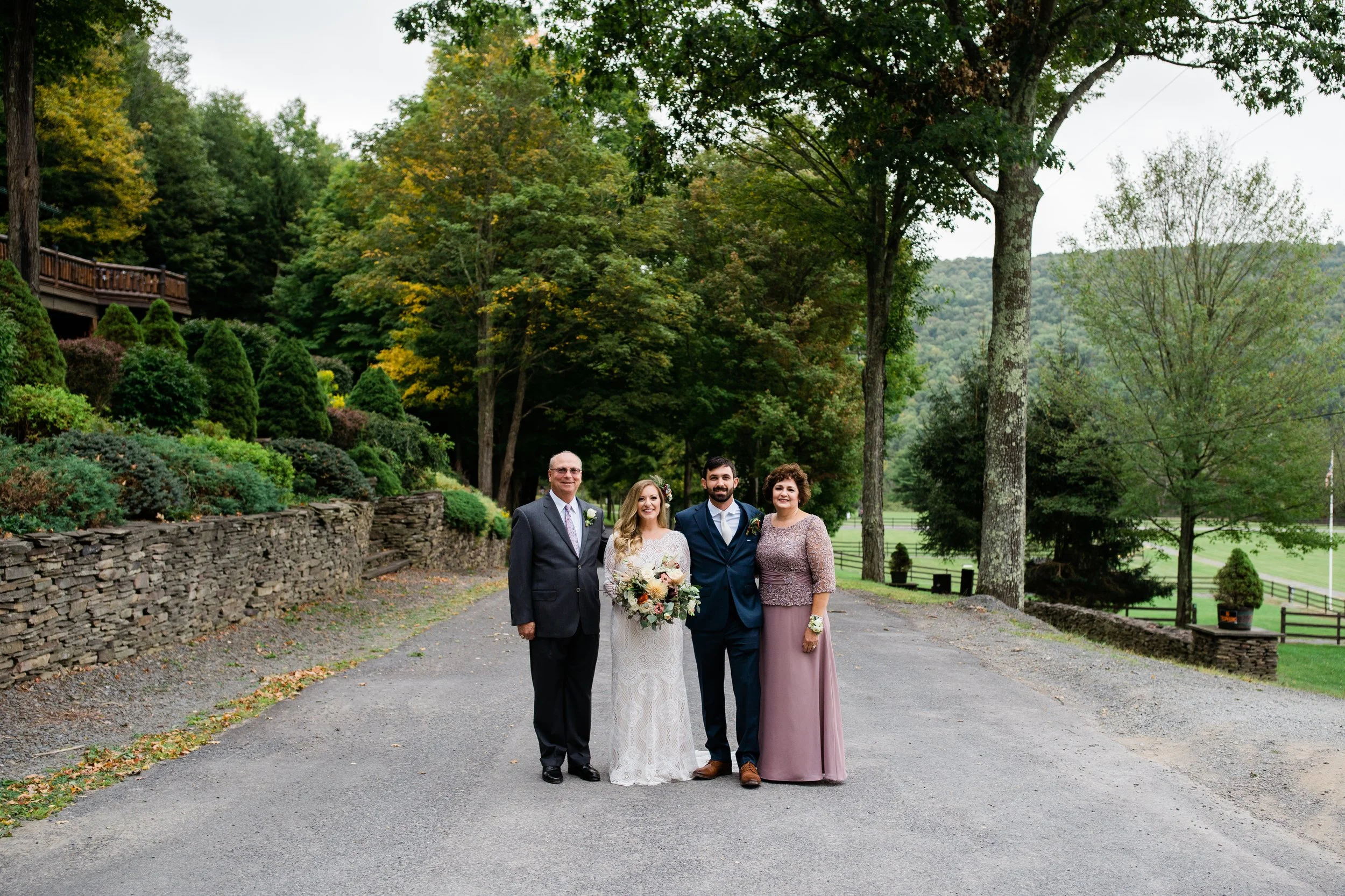 Four people, including a bride in a white lace wedding dress holding a bouquet and three others in formal attire, standing on a gravel path in a lush, green outdoor setting with tall trees and a stone wall.