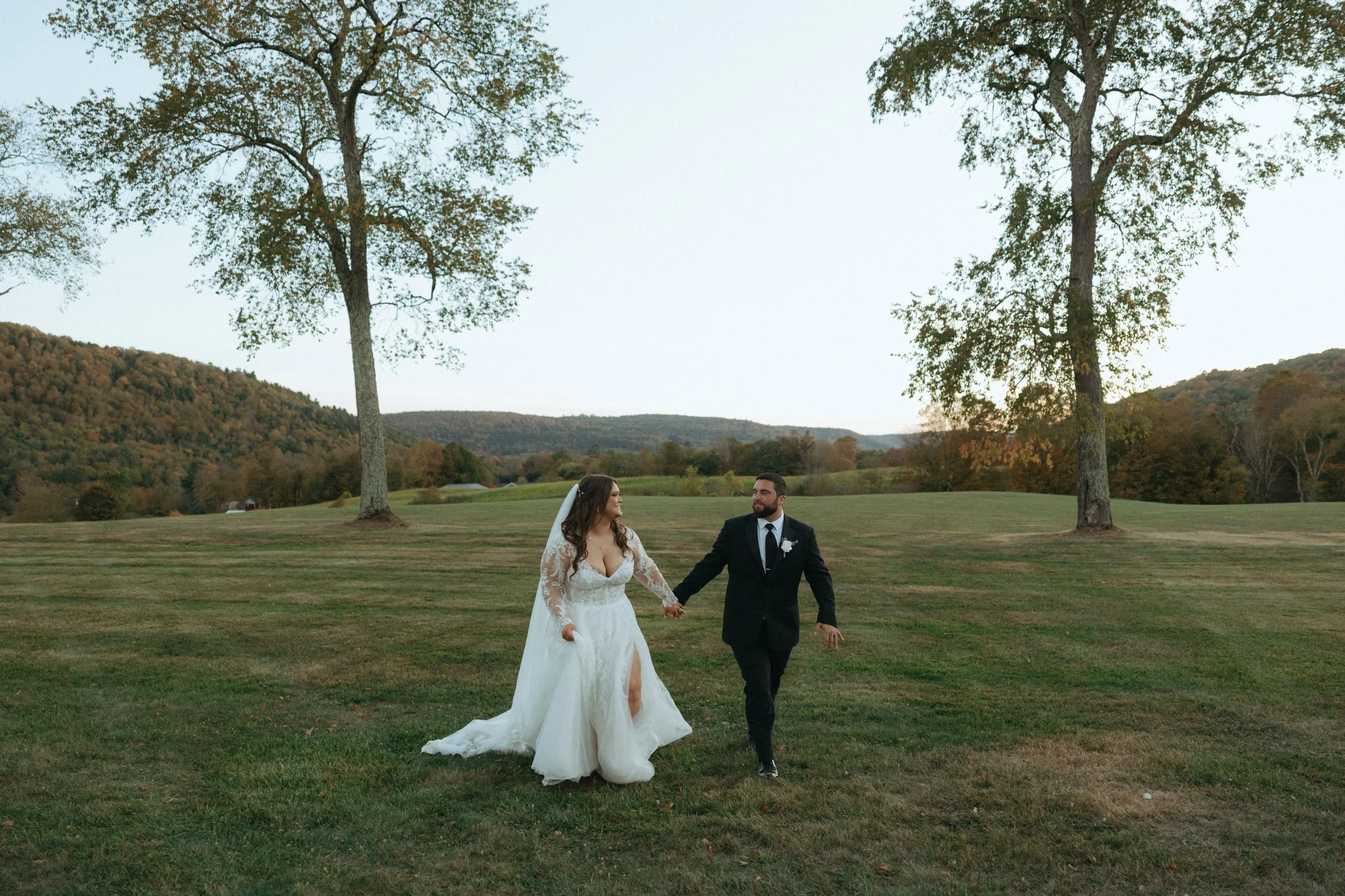 A bride and groom holding hands and walking on a grassy field with trees and hills in the background during an outdoor wedding.