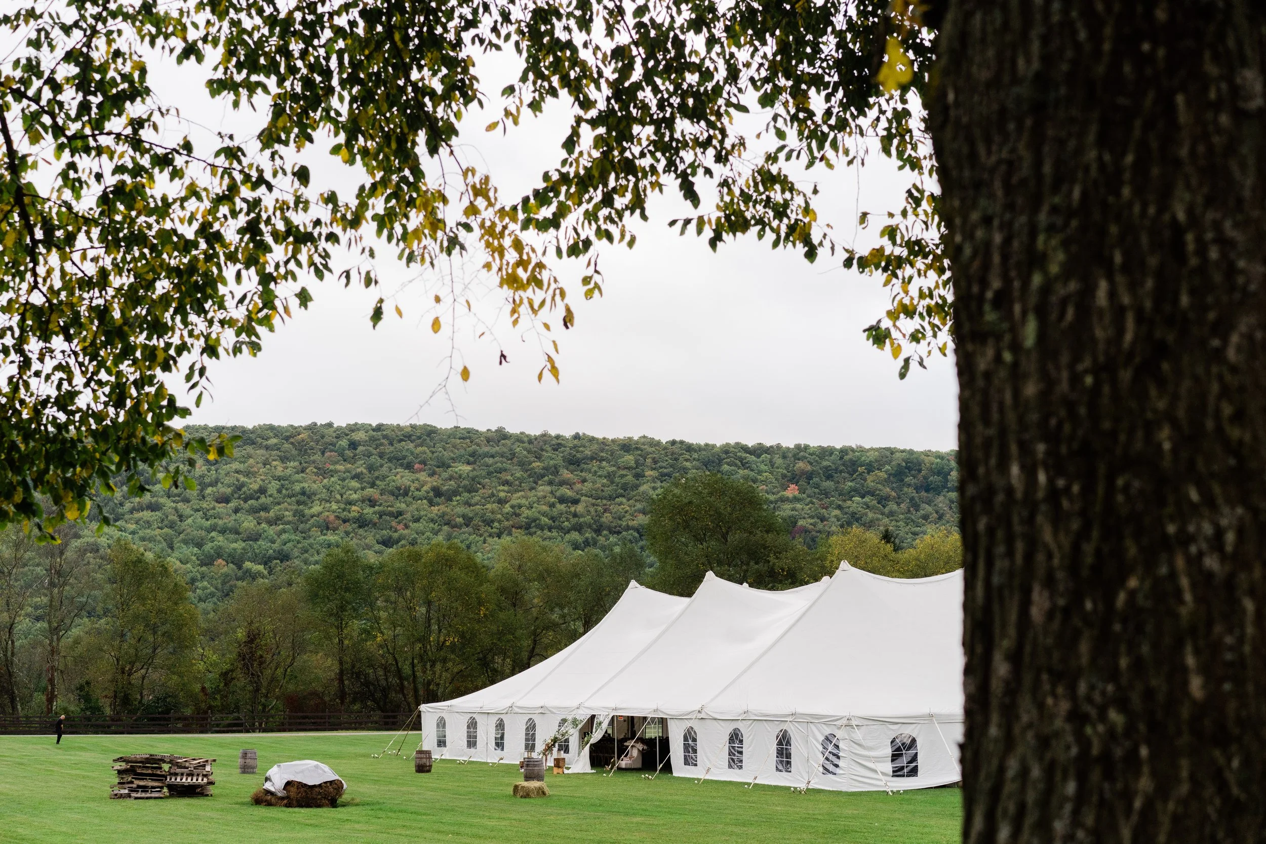 Large white event tents set up outdoors on green grass, with a backdrop of a hilly forested landscape and a cloudy sky.