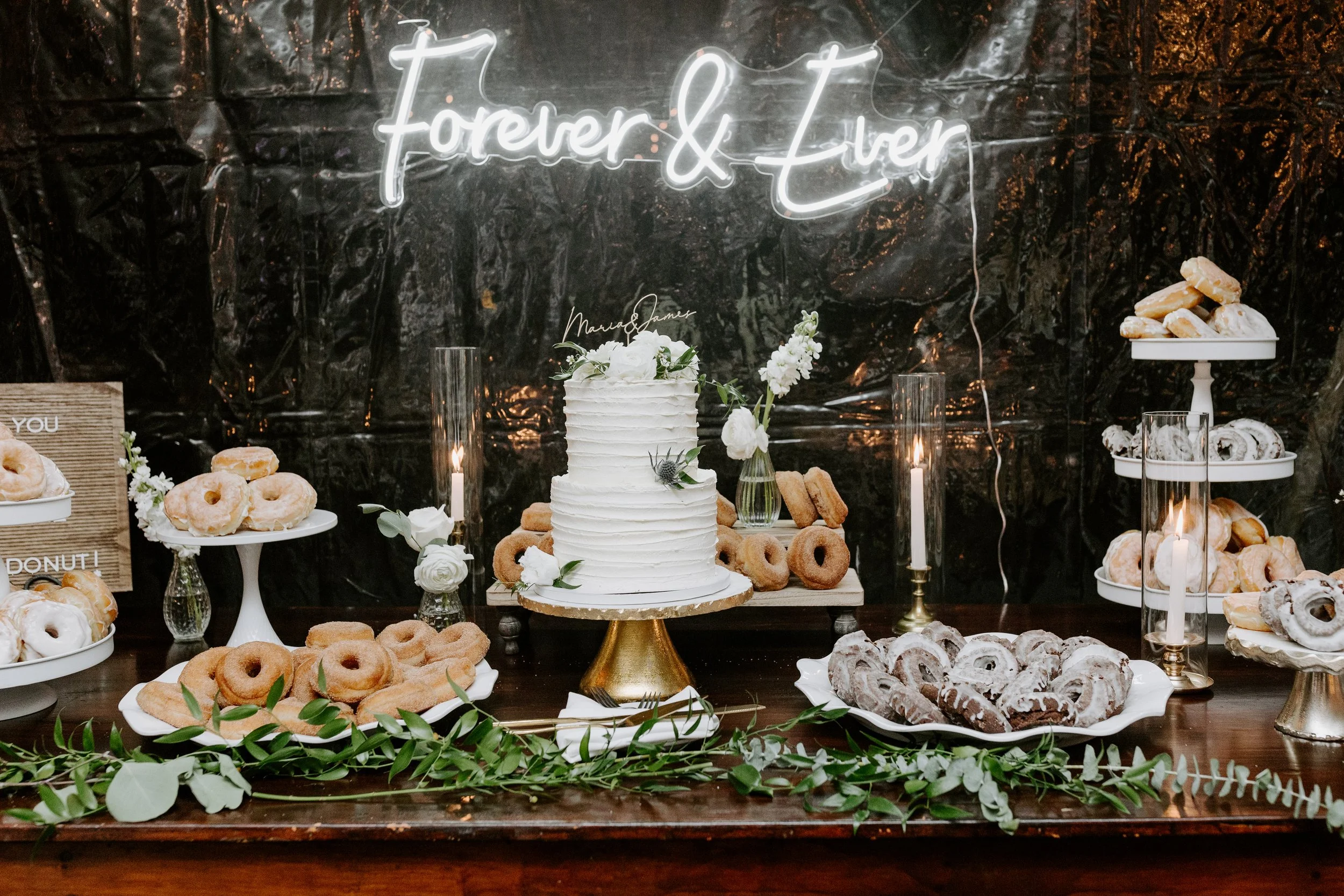 A dessert table with a white frosted cake, various donuts, cookies, and pastries displayed on white tiers and plates, decorated with flowers and greenery, with a "Forever & Ever" neon sign in the background.