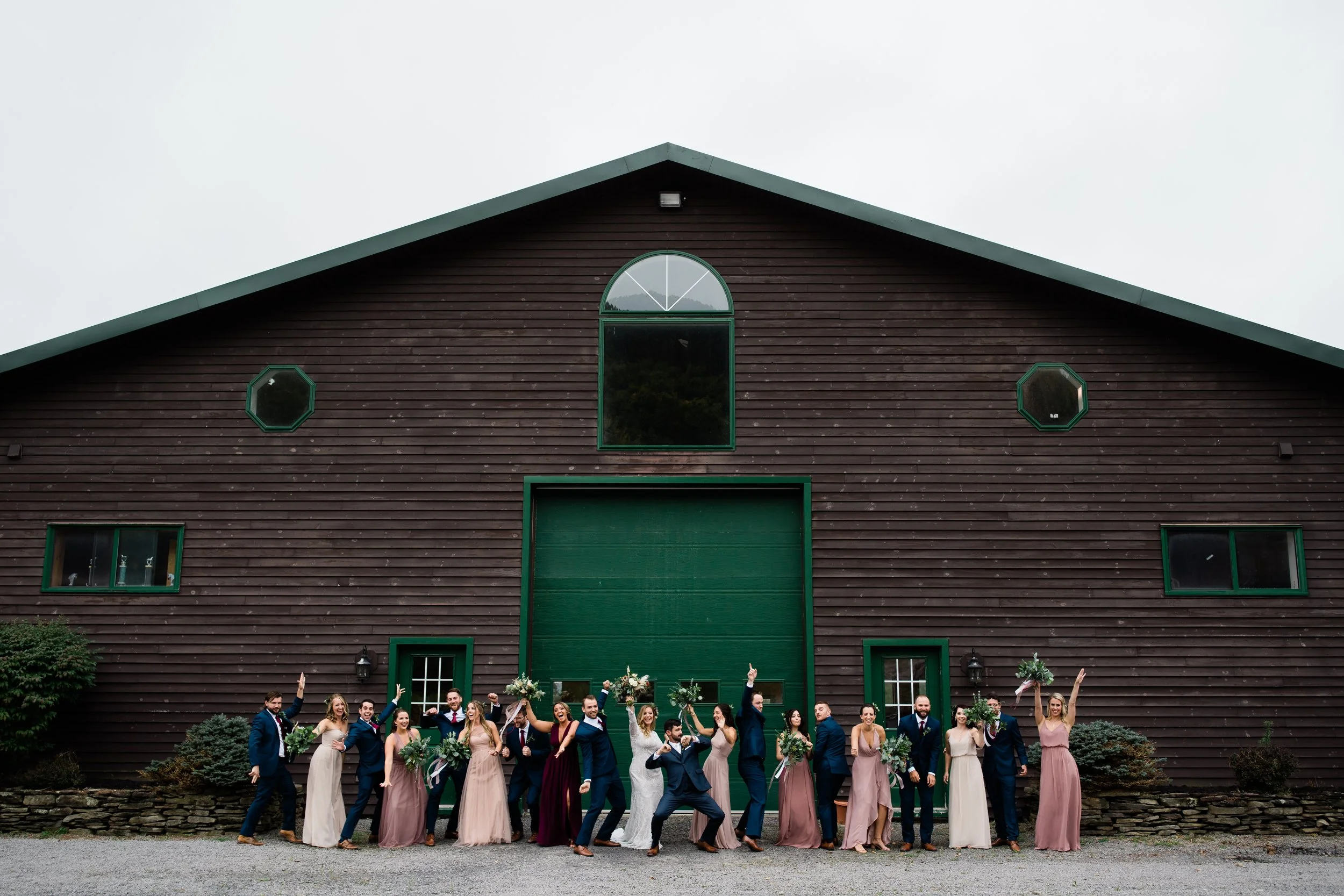 A large group of wedding party members in formal attire, holding bouquets, standing in front of a rustic wooden building with green accents, celebrating outdoors.