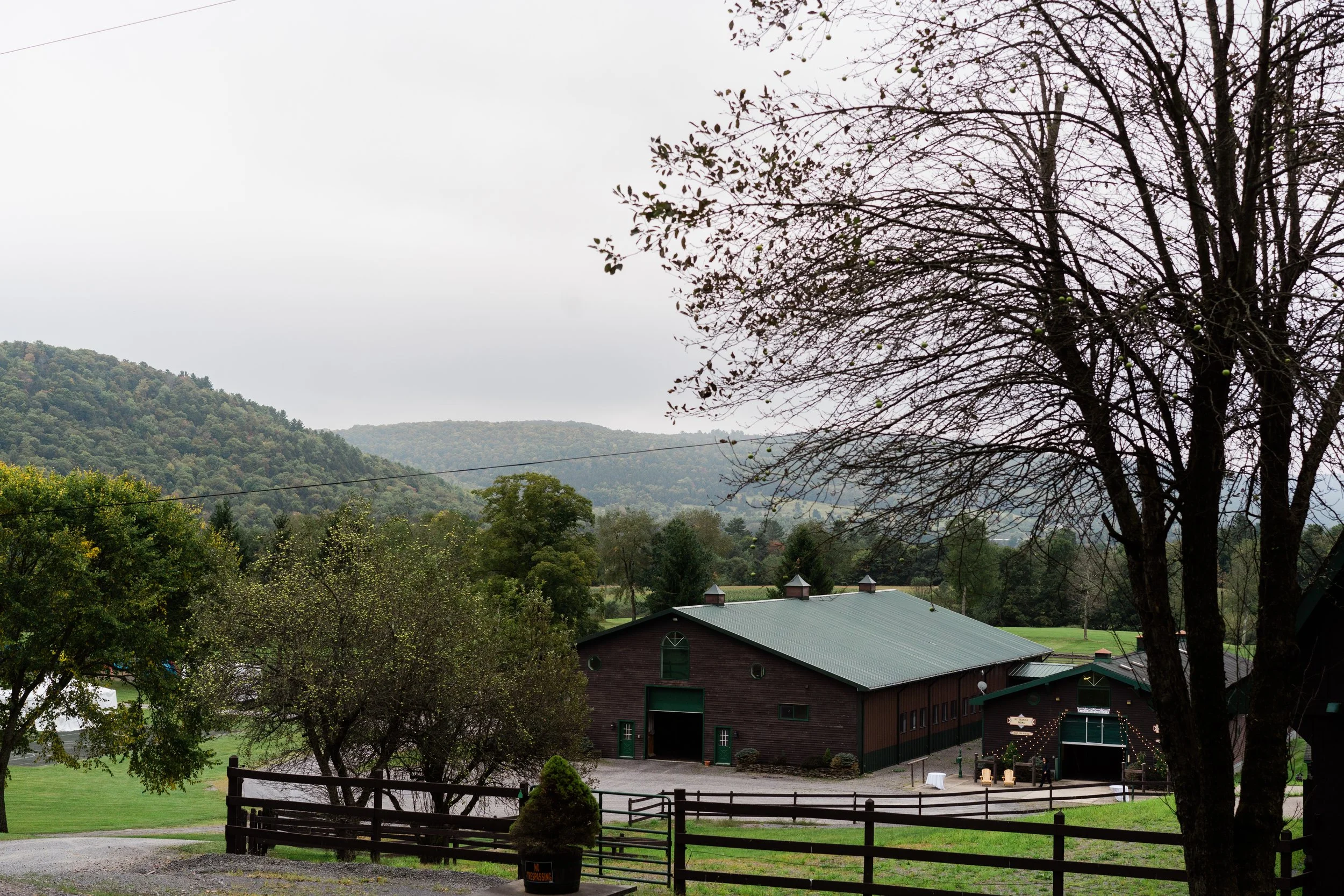 A rural landscape with a barn, trees, and a hilly background under an overcast sky.