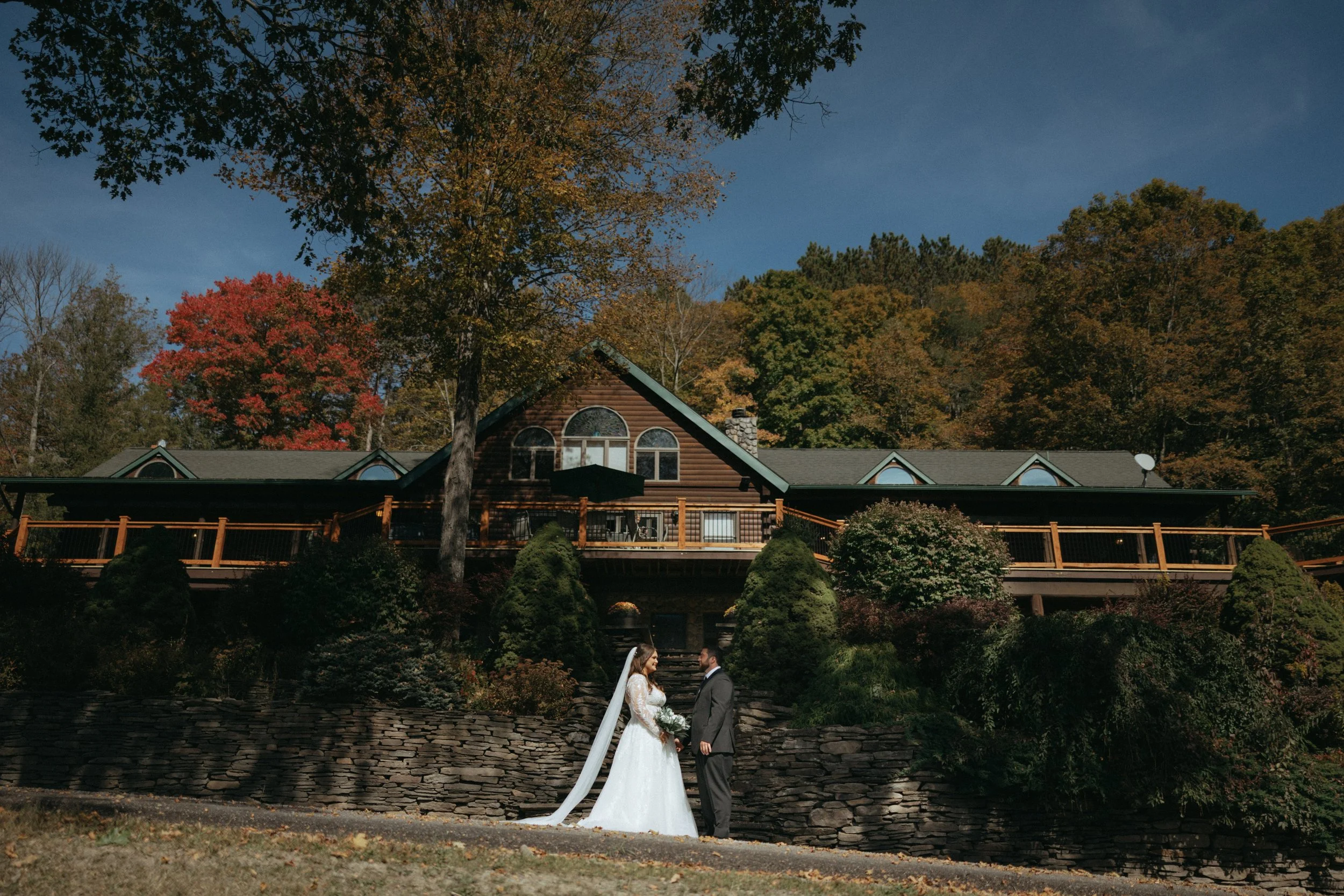 A bride and groom standing on a stone pathway outdoors during daytime, holding hands and gazing at each other, with a two-story house and colorful autumn trees in the background.