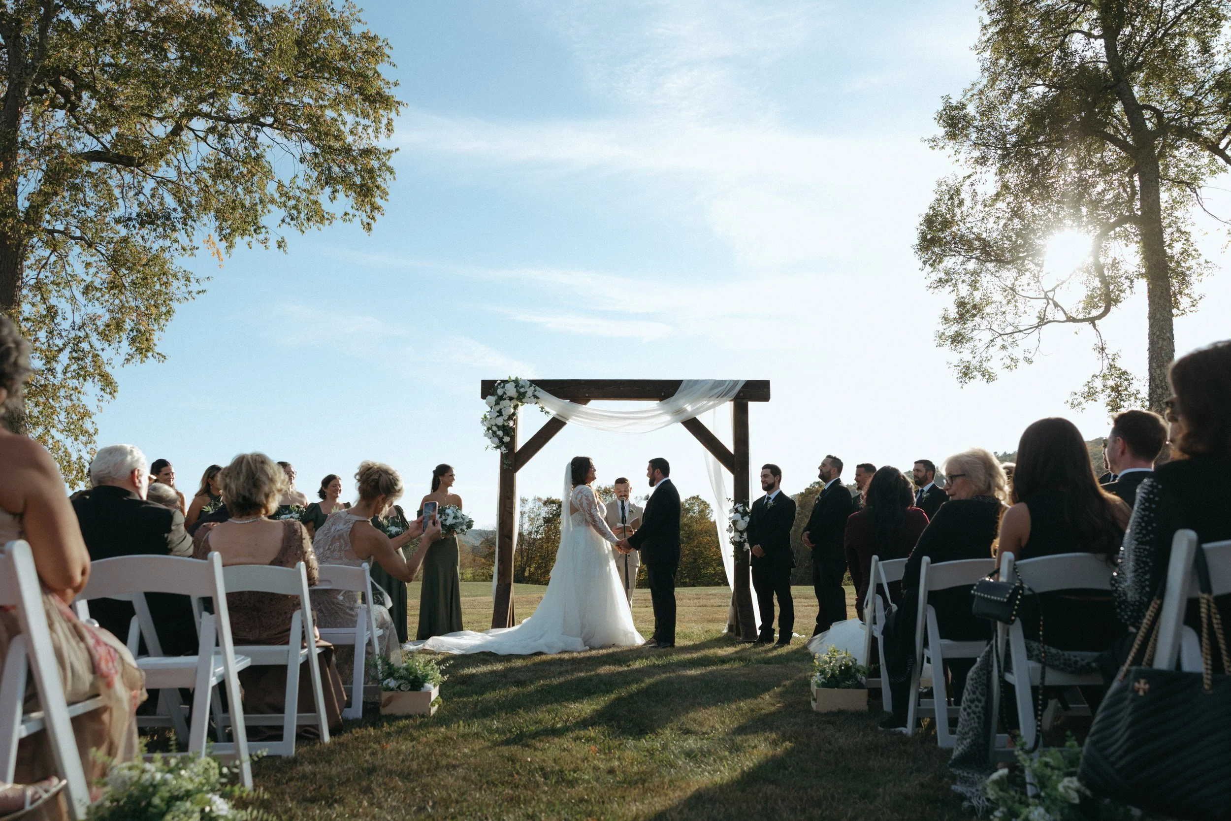 Outdoor wedding ceremony with a bride and groom exchanging vows under a wooden arch decorated with flowers, surrounded by seated guests on a grassy field, with trees and the sun shining in the background.
