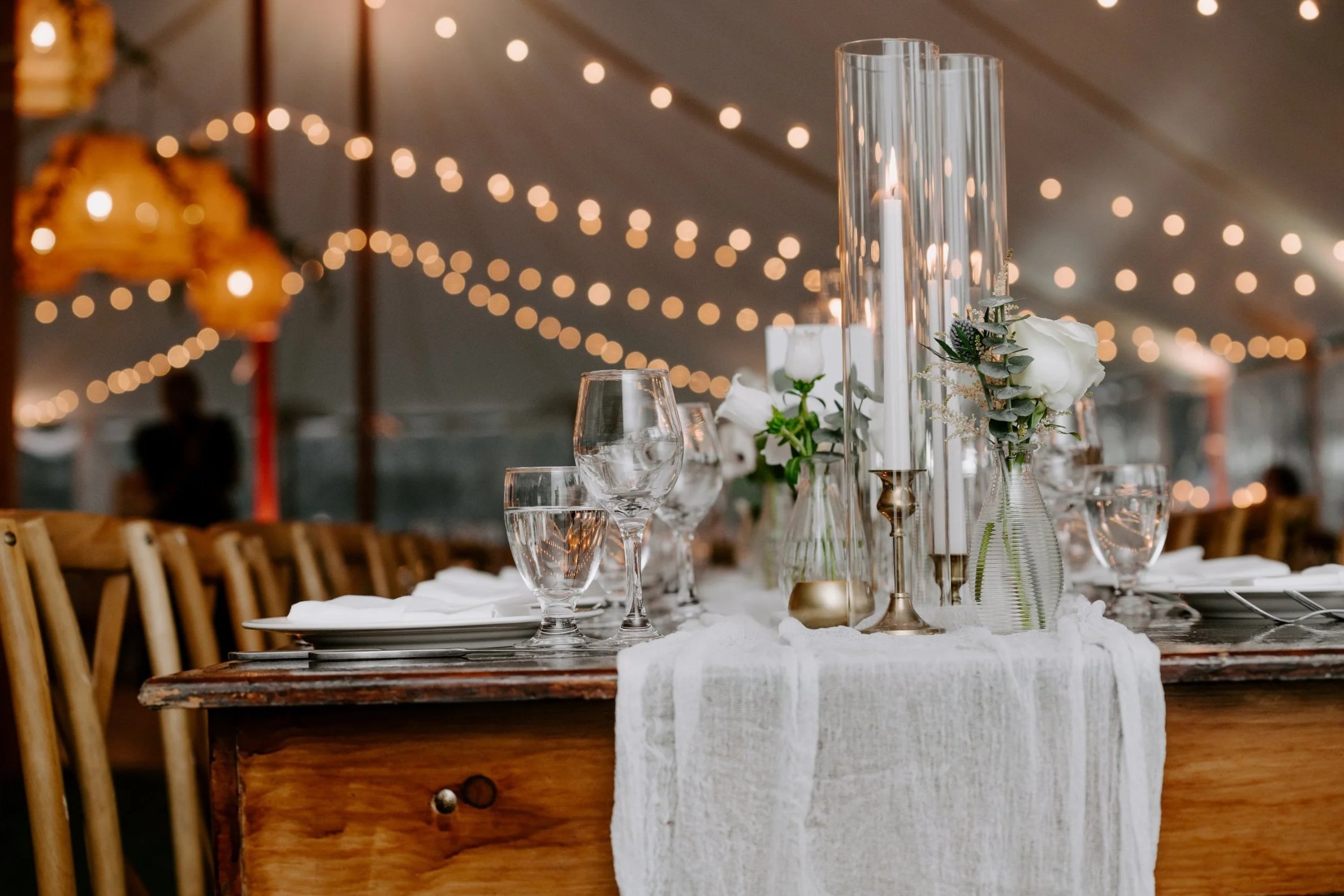 A table decorated for a formal event with white flowers, candles in tall glass holders, and multiple wine glasses, set under string lights and lanterns in a tent.
