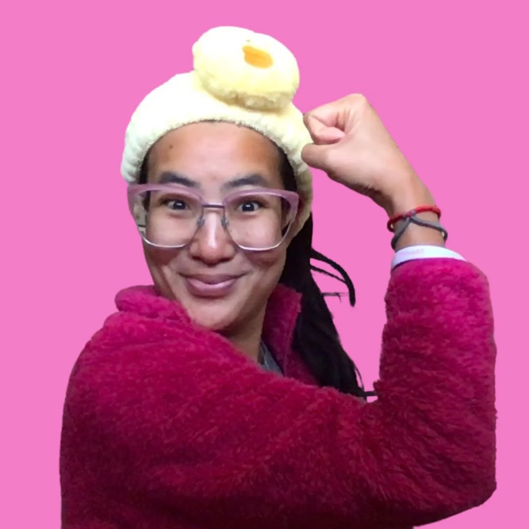 Kyla Mitsunaga smiling in a powerful Rosie the Riveter pose, wearing a pink fuzzy sweater and an iconic yellow chick headband against a solid pink background.