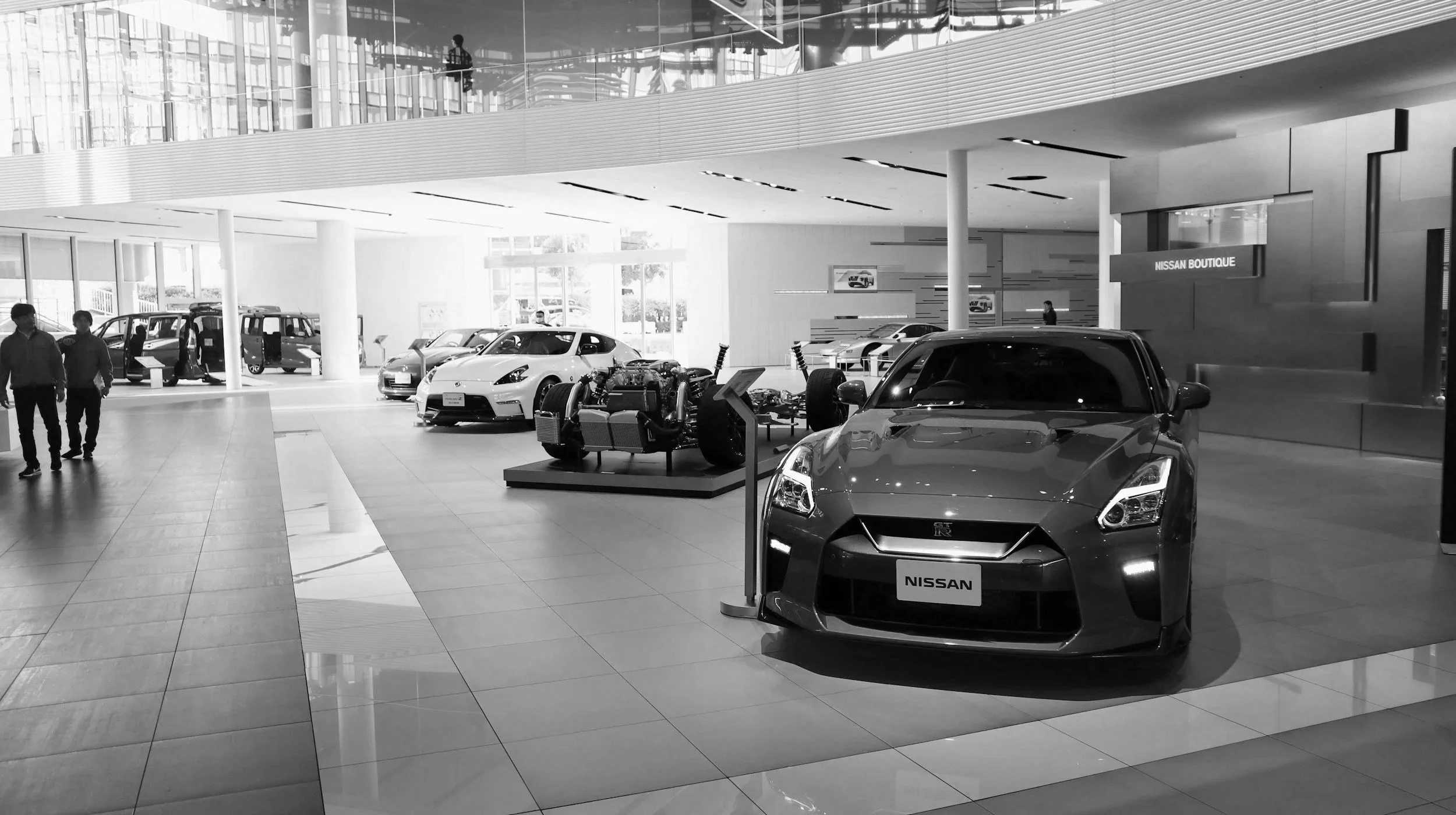 Interior of a Nissan car dealership showroom featuring various Nissan cars, including a sports car at the front, with a modern design and large glass windows, two people walking, and a Nissan Boutique sign.