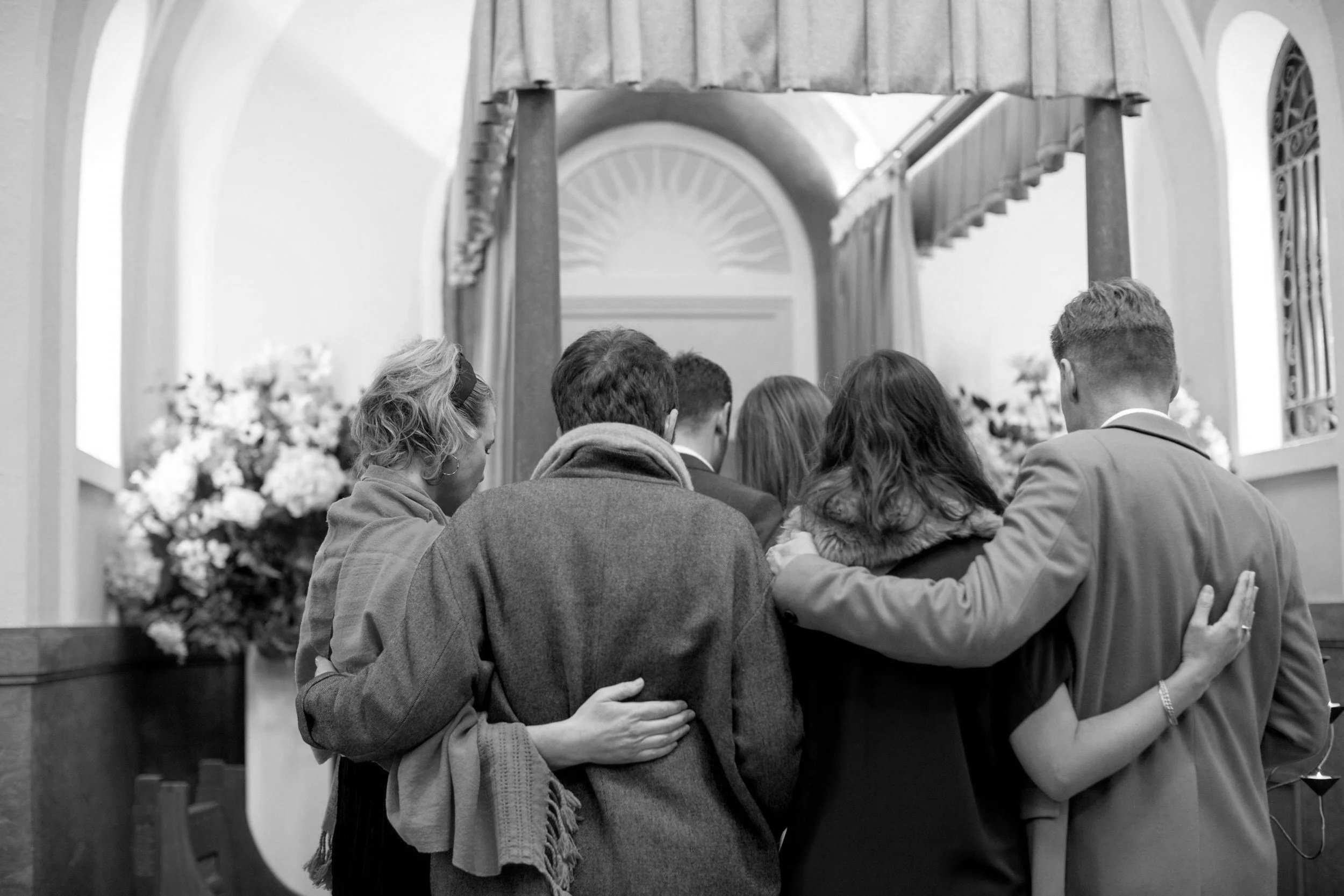 A group of people standing close together, facing away, with their arms around each other, in a church or similar setting, with a cross and flowers visible in the background.