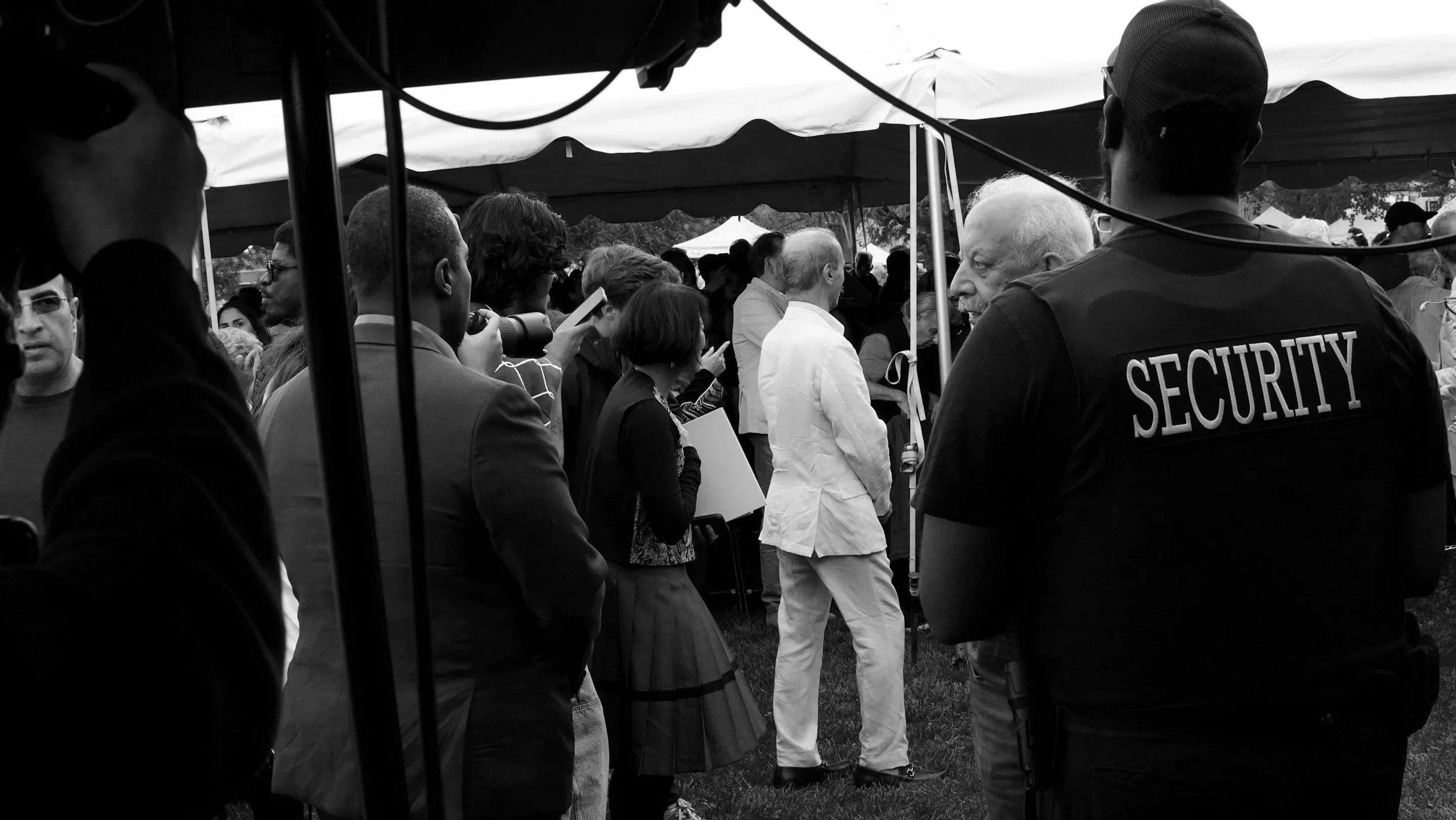 Crowd of people gathered under a large tent, with security personnel in the foreground.