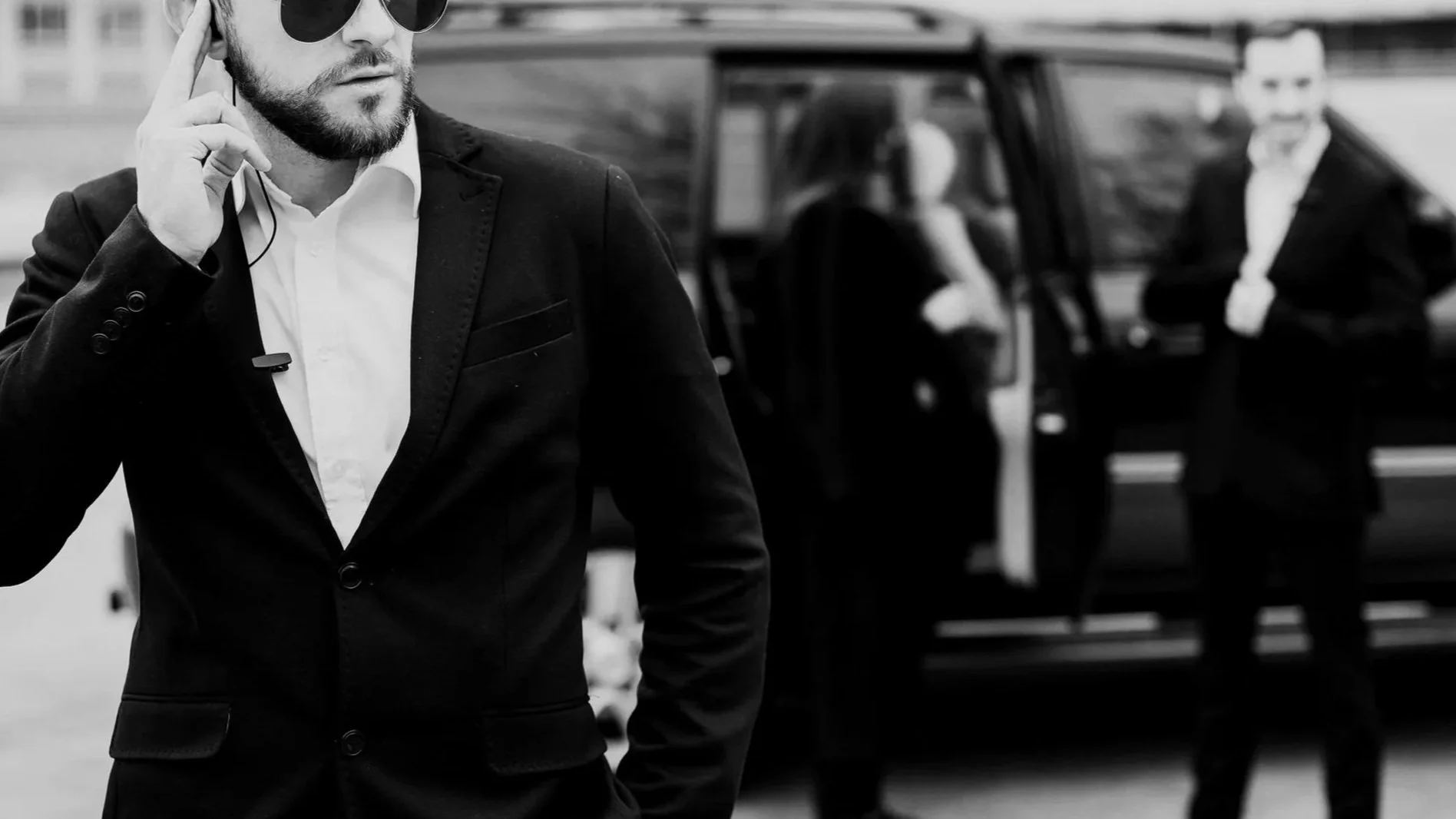 A man in a suit and sunglasses talking on a cellphone in front of a black SUV, with a woman and another man in business attire standing beside the vehicle in the background.