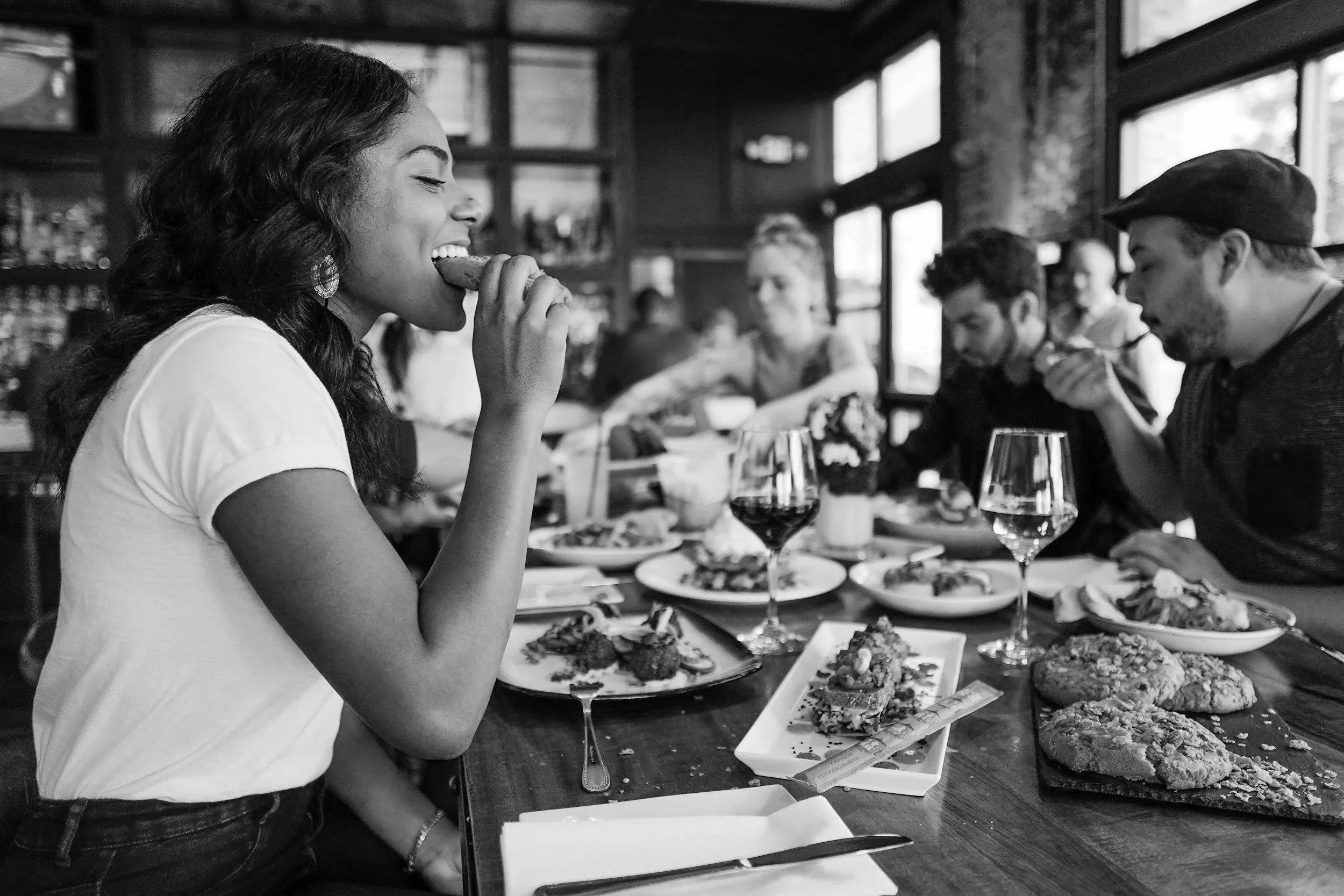 A woman with dark, wavy hair is taking a bite of a taco at a restaurant table filled with various plates of food, including other tacos and appetizers. In the background, other diners are seated, and there are large windows letting in natural light.