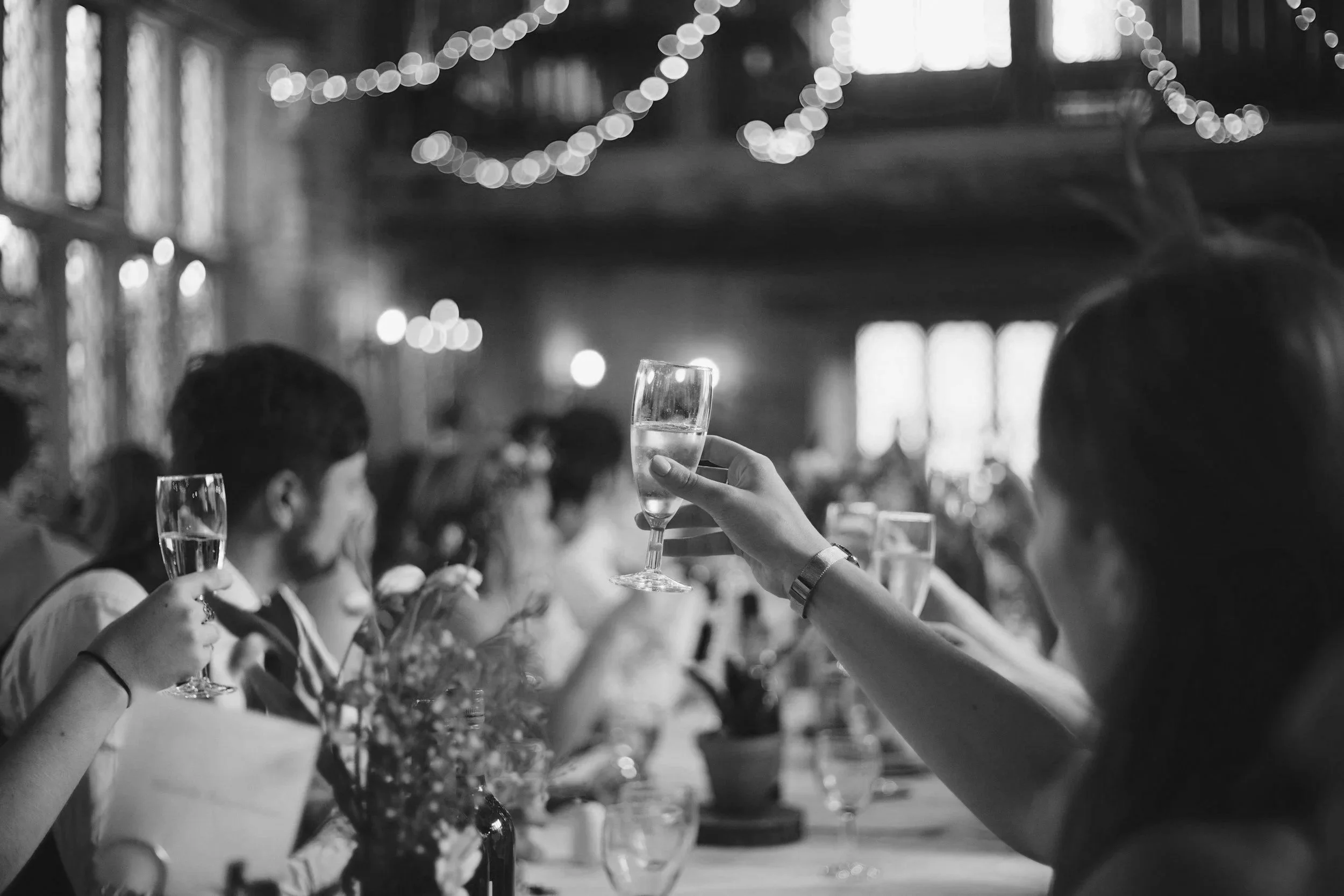 People celebrating, raising champagne glasses, in a decorated social setting with string lights and large windows.