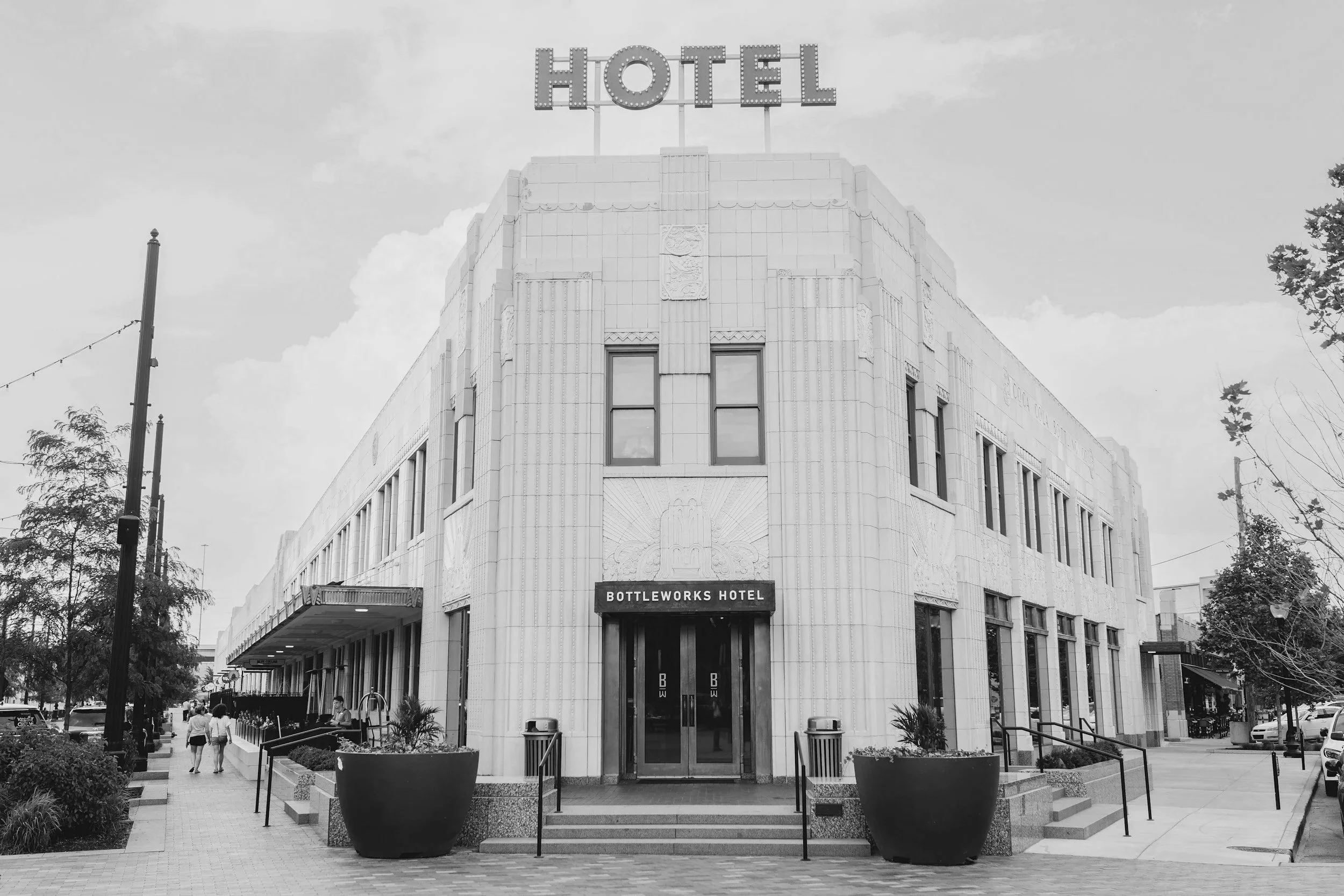 Front view of the Bottleworks Hotel, a historic building with art deco architecture, featuring a sign on top that reads 'HOTEL', and a sign below that says 'Bottleworks Hotel'. There are people walking on the sidewalk and trees on either side of the entrance.