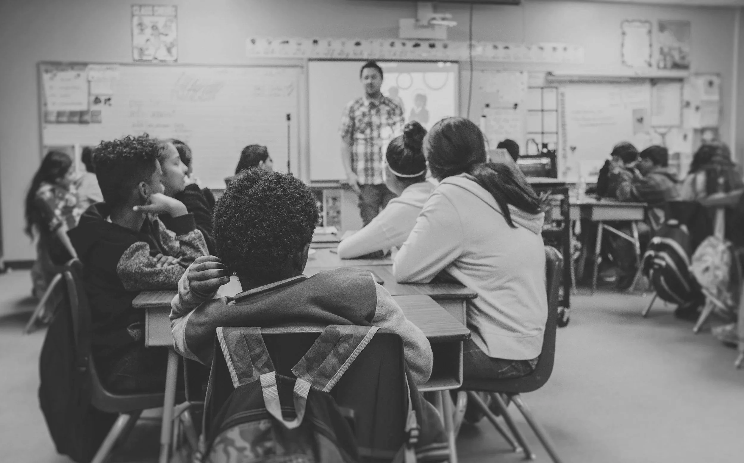 A classroom with students sitting at desks, listening to a teacher at the front. The teacher stands near a whiteboard and a projector screen, with educational posters on the walls.