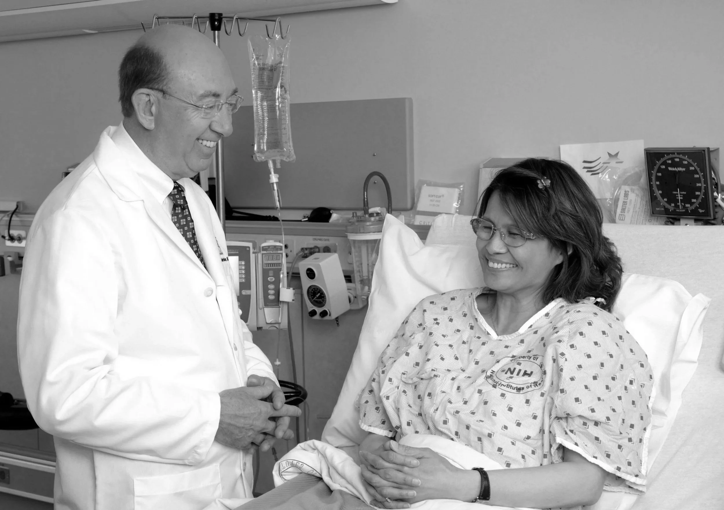 A doctor is smiling and holding the hand of a patient who is lying in a hospital bed. The patient is also smiling. Medical equipment can be seen in the background.