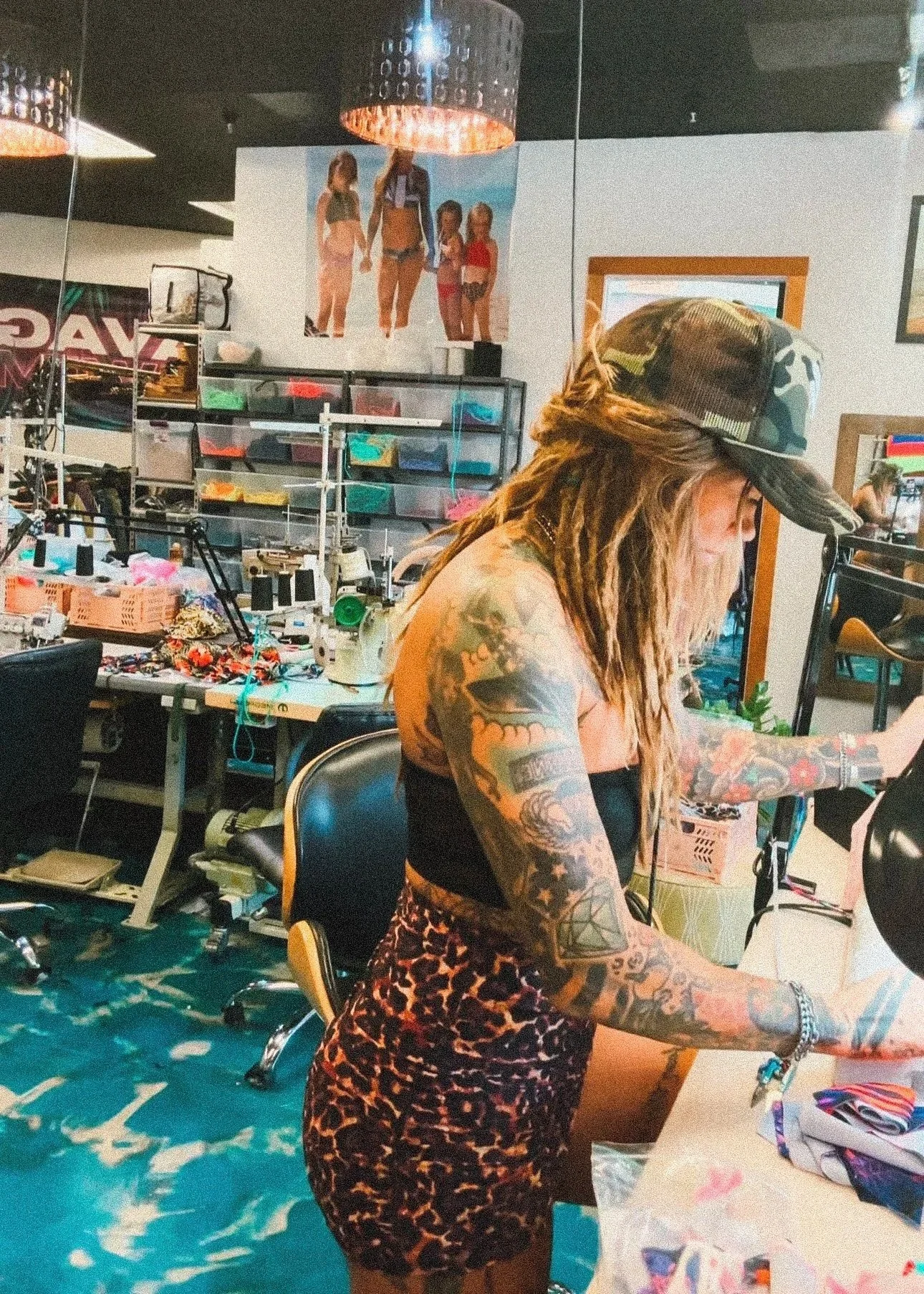 A woman with extensive tattoos, wearing a camouflage cap and leopard print skirt, working at a crafting table in a tattoo studio. The background shows shelves with supplies, a mirror on the wall, and a poster of four women in swimsuits on the wall.