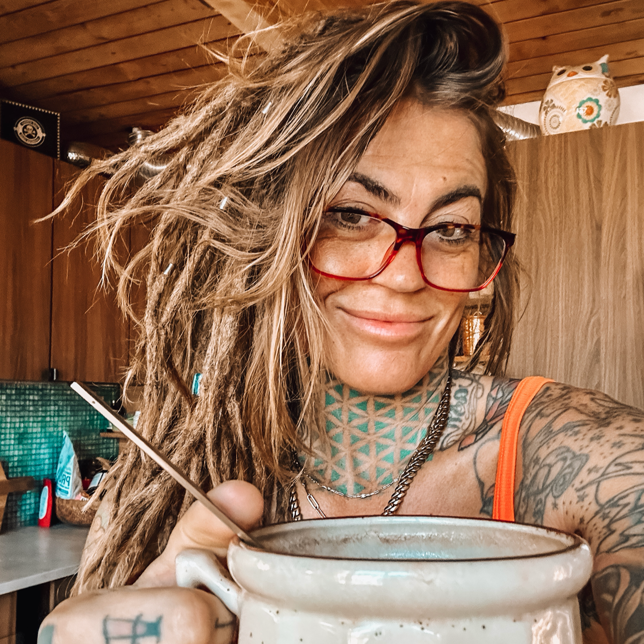A woman with tattoos and dreadlocks, wearing red glasses and an orange tank top, smiling while holding a mug.